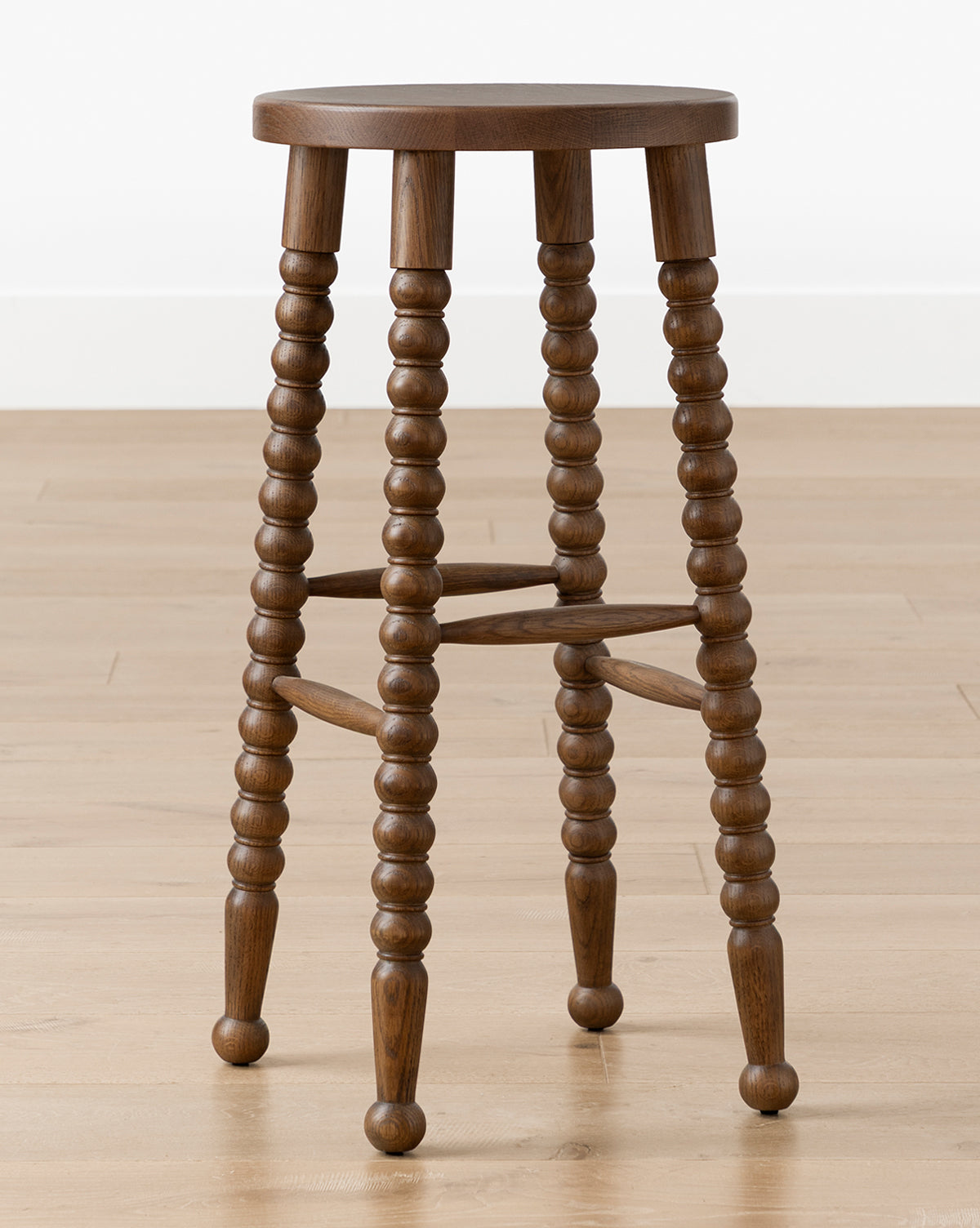 The Makerspalm Heathcliff Stool features a walnut oak finish, four turned legs with crossbars, and a round wooden seat, shown on a light wood floor against a plain white wall. McGee & Co.
