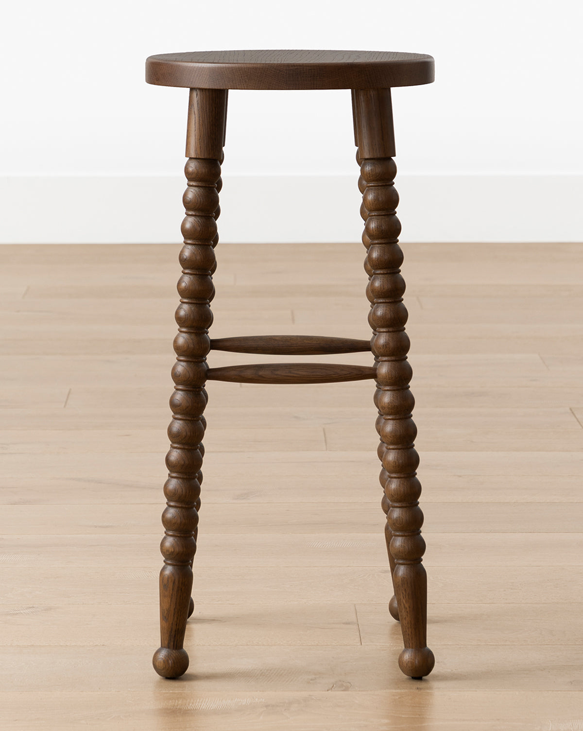 The Makerspalm Heathcliff Stool features a dark walnut oak finish, three intricately turned legs, and a round seat, shown on a light wood floor against a white wall. McGee & Co.