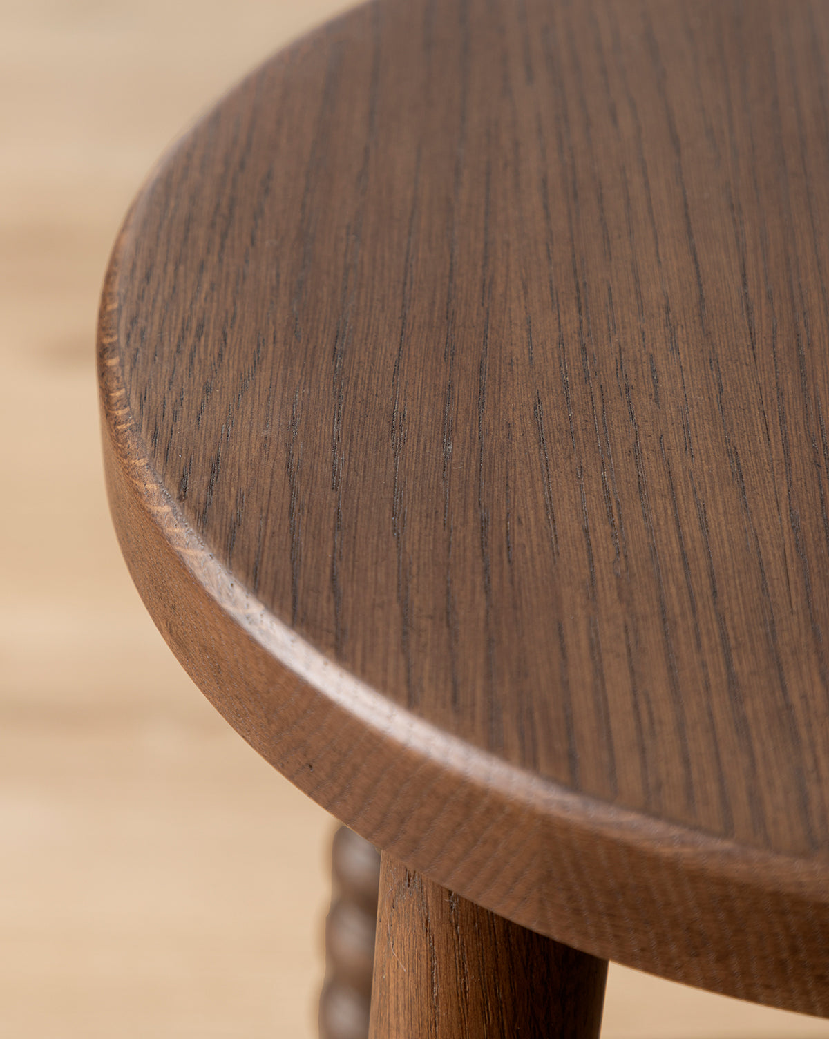 Close-up of a Makerspalm Heathcliff Stool shows its round tabletop with a smooth, dark brown walnut oak finish and visible wood grain, plus one distinctive bobbin-style leg—finished in the refined style of McGee & Co.