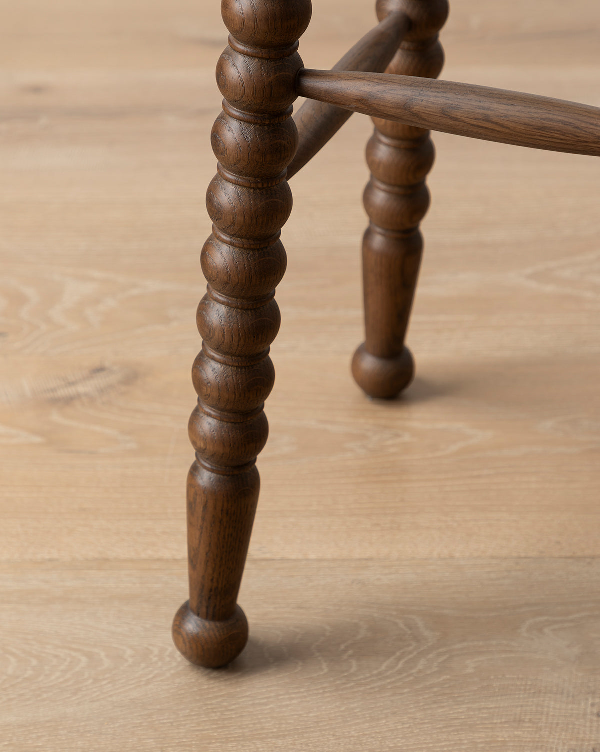 Close-up of a Makerspalm Heathcliff Stool leg with rounded, bobbin-style segments in a walnut oak finish, highlighting the detailed design and connecting rod against a light wood floor—McGee & Co.