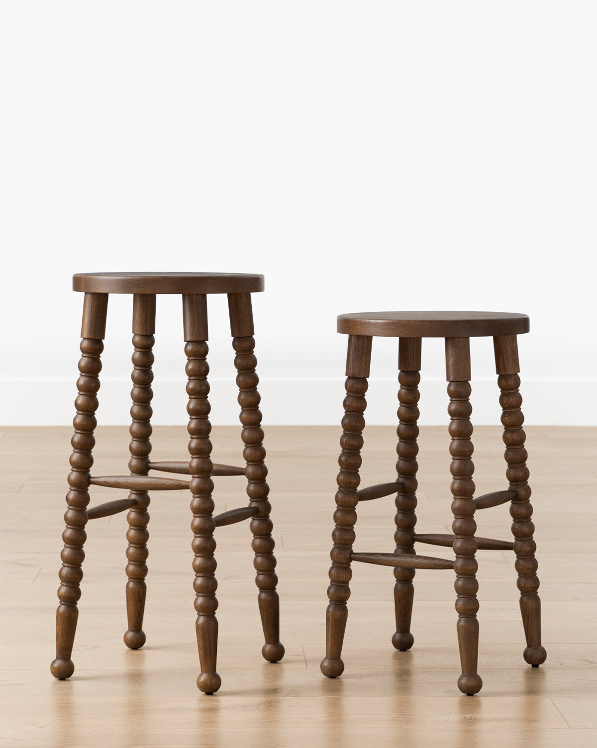 Two Makerspalm Heathcliff Stools with round seats and bobbin-style legs in walnut oak finish are set on a light wood floor against a plain white wall, McGee & Co.