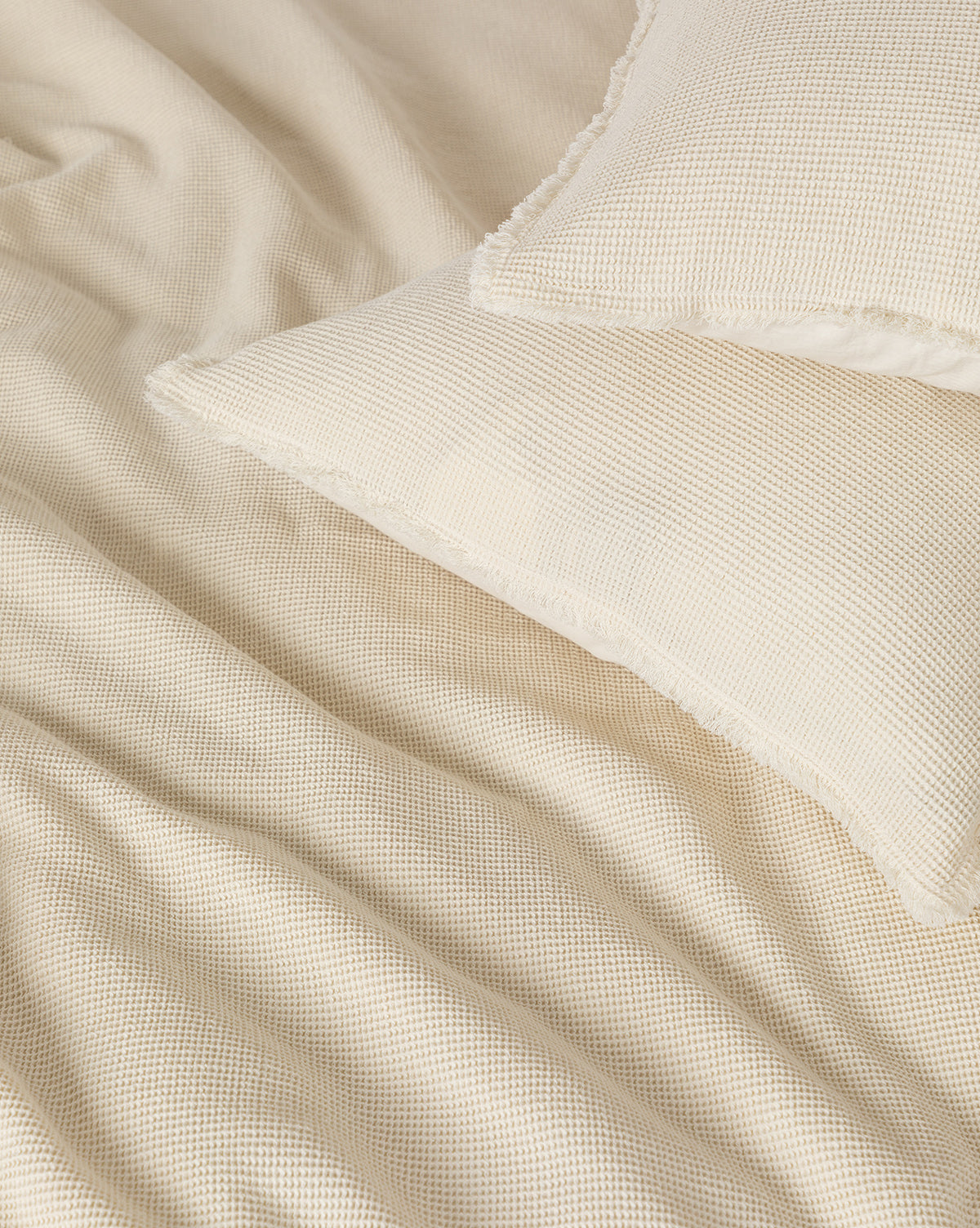 Close-up of two Helene Waffle Shams (Set of 2) in cream-colored cotton with a textured waffle weave, paired with a matching bedspread. Gentle fabric folds enhance the cozy, soothing appeal of these neutral-toned shams.