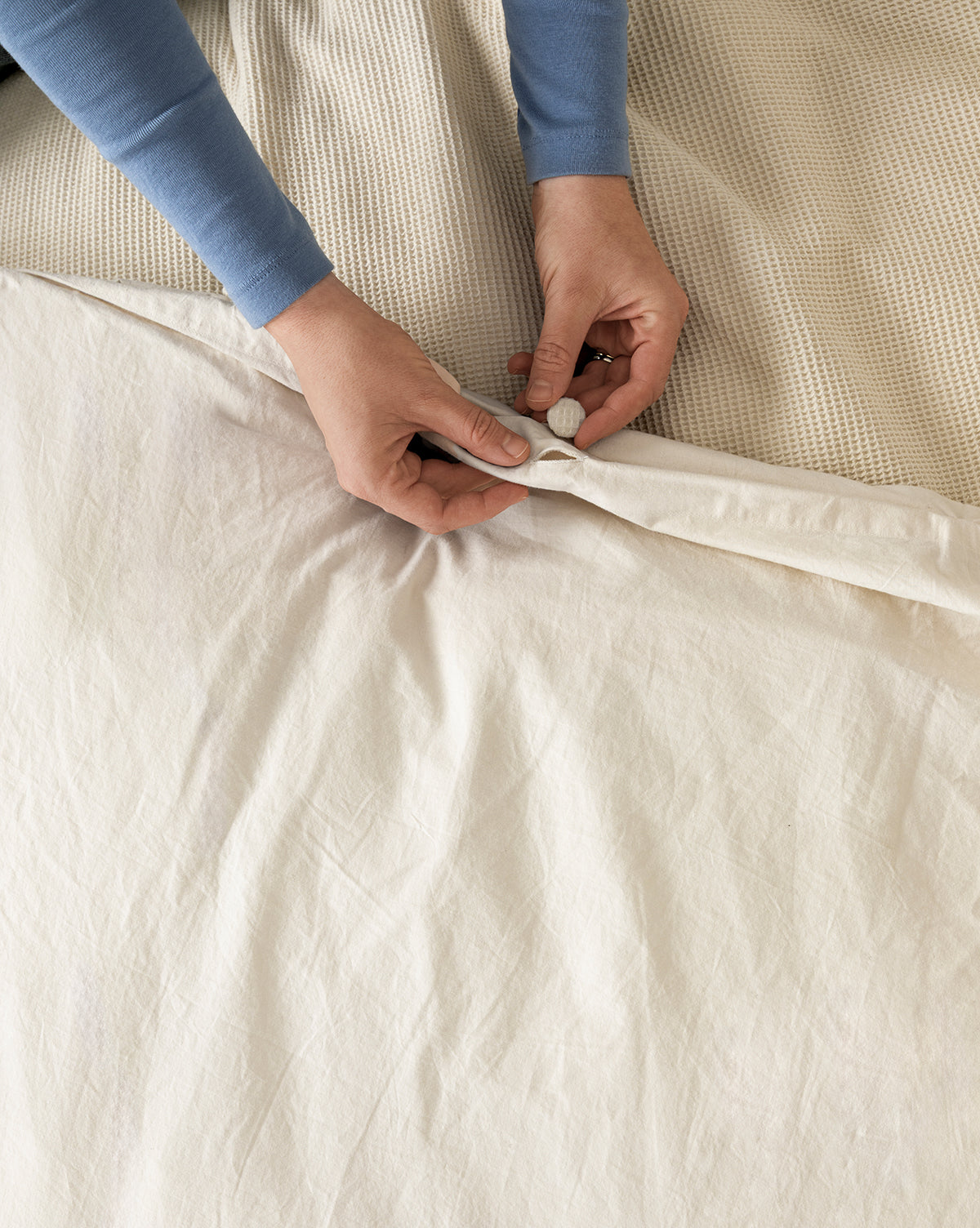 A person in a blue long-sleeve shirt fastens a button on the Helene Waffle Duvet Cover over textured bedding, giving the perfect finishing touch to their cozy bed.