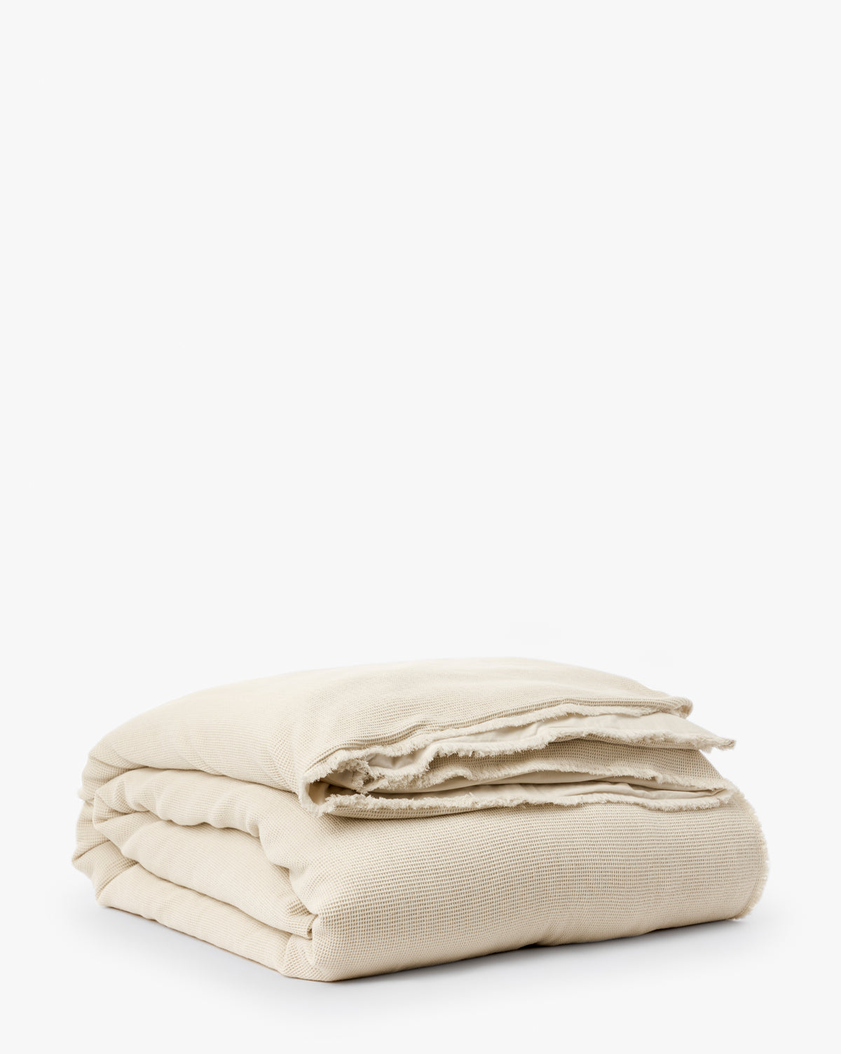 The Helene Waffle Duvet Cover, neatly folded in off-white cotton with frayed edges, adds charm and depth to textured bedding, shown against a plain white background.