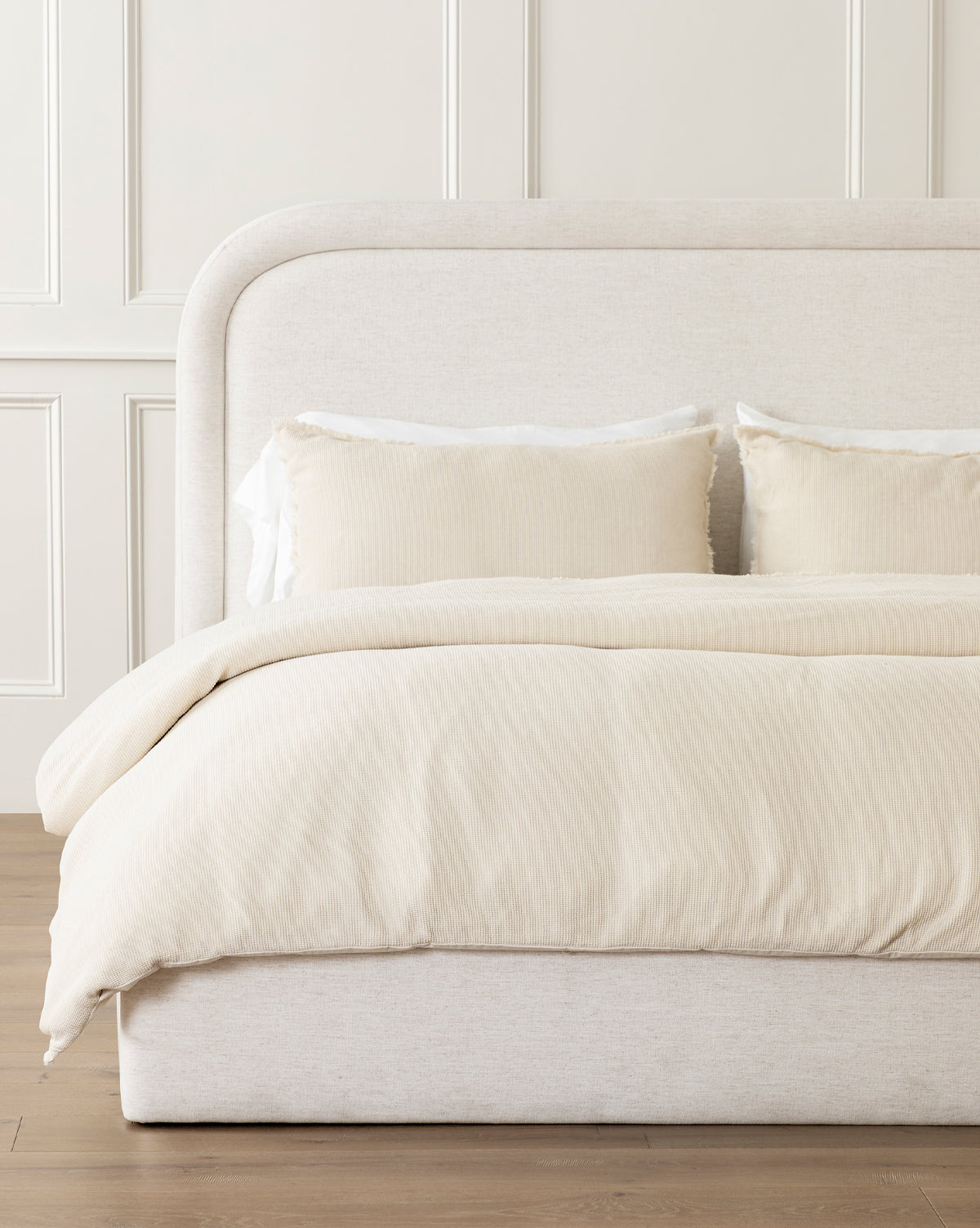 A neatly made bed features the beige upholstered headboard and Helene Waffle Duvet Cover in light cream, with matching textured pillows—all set against a white paneled wall on a wooden floor.