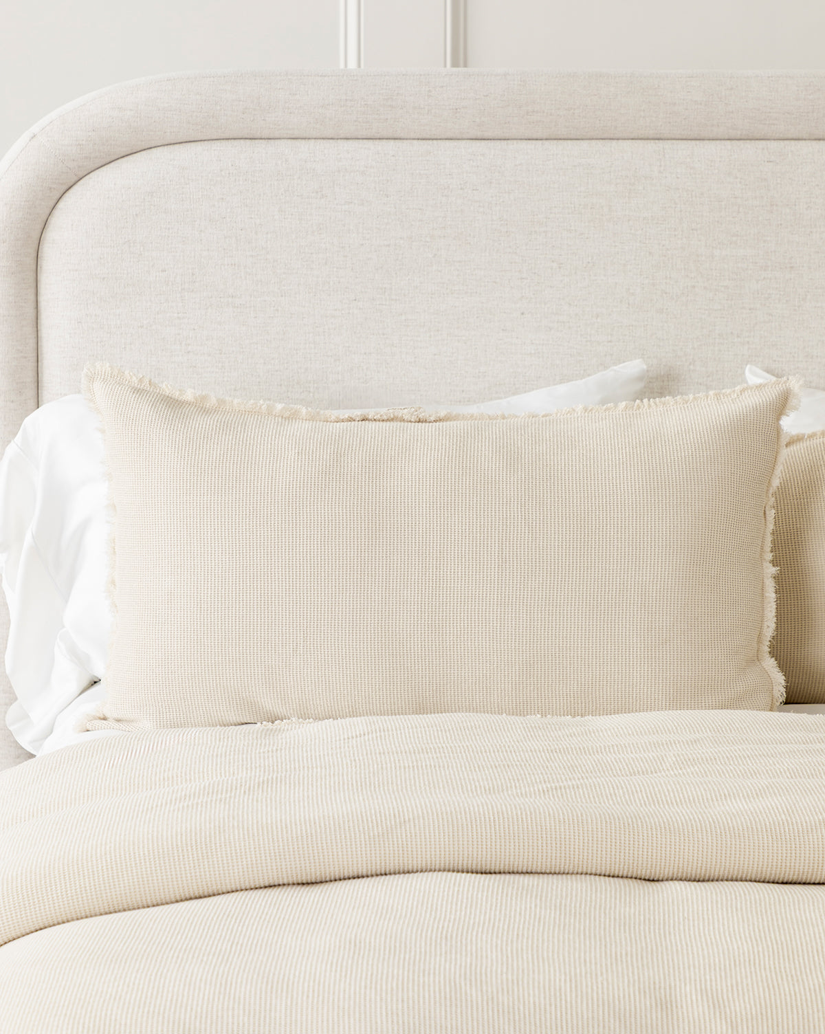 A neatly made bed features the calming neutral tone of Helene Waffle Shams (Set of 2), white pillows, a textured cream pillow with fringed edges, and a beige upholstered headboard for a serene, inviting setting.