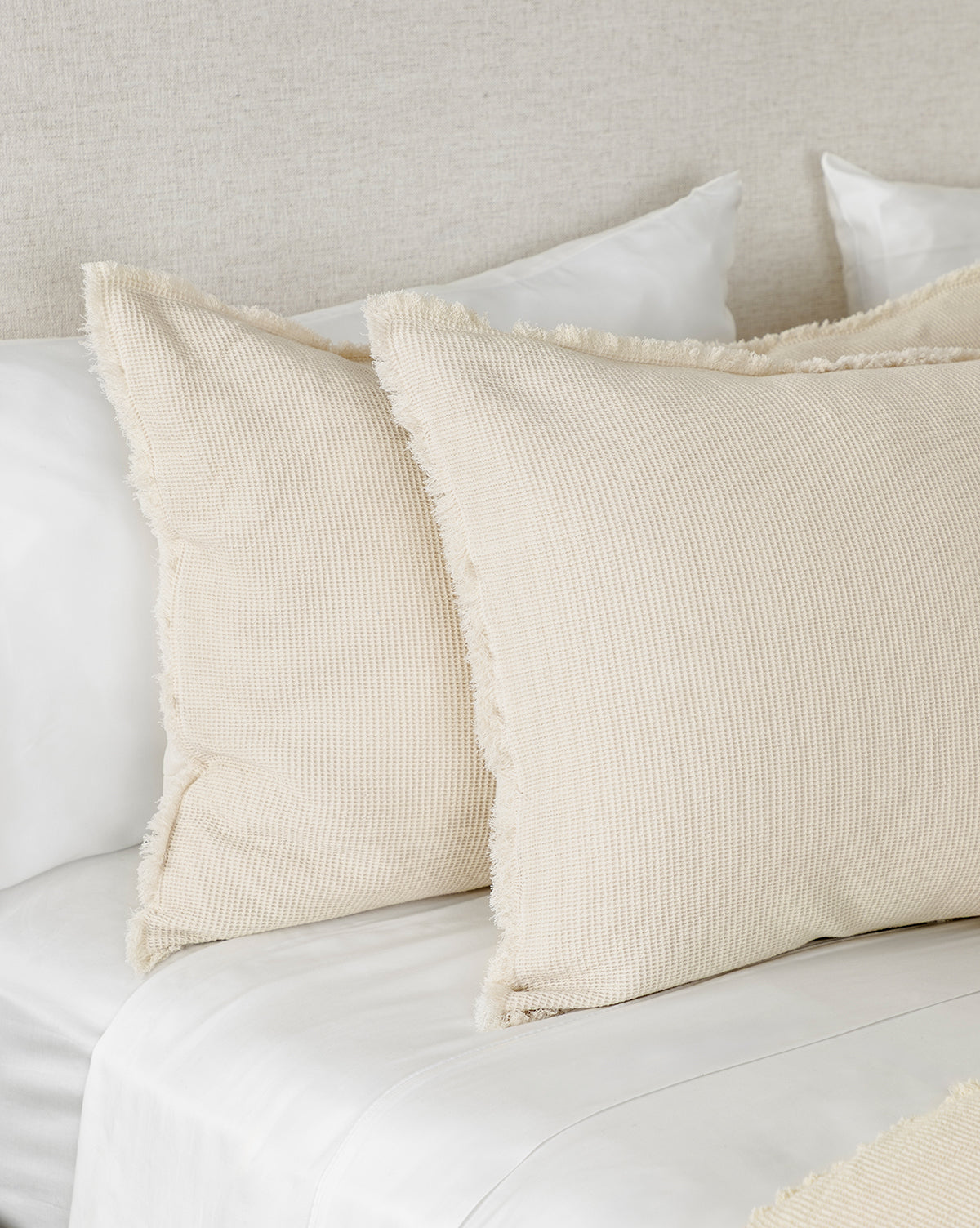 Two beige textured pillows with fringed edges rest on a neatly made bed with white sheets, complemented by Helene Waffle Shams (Set of 2) in a neutral tone against a light fabric headboard.