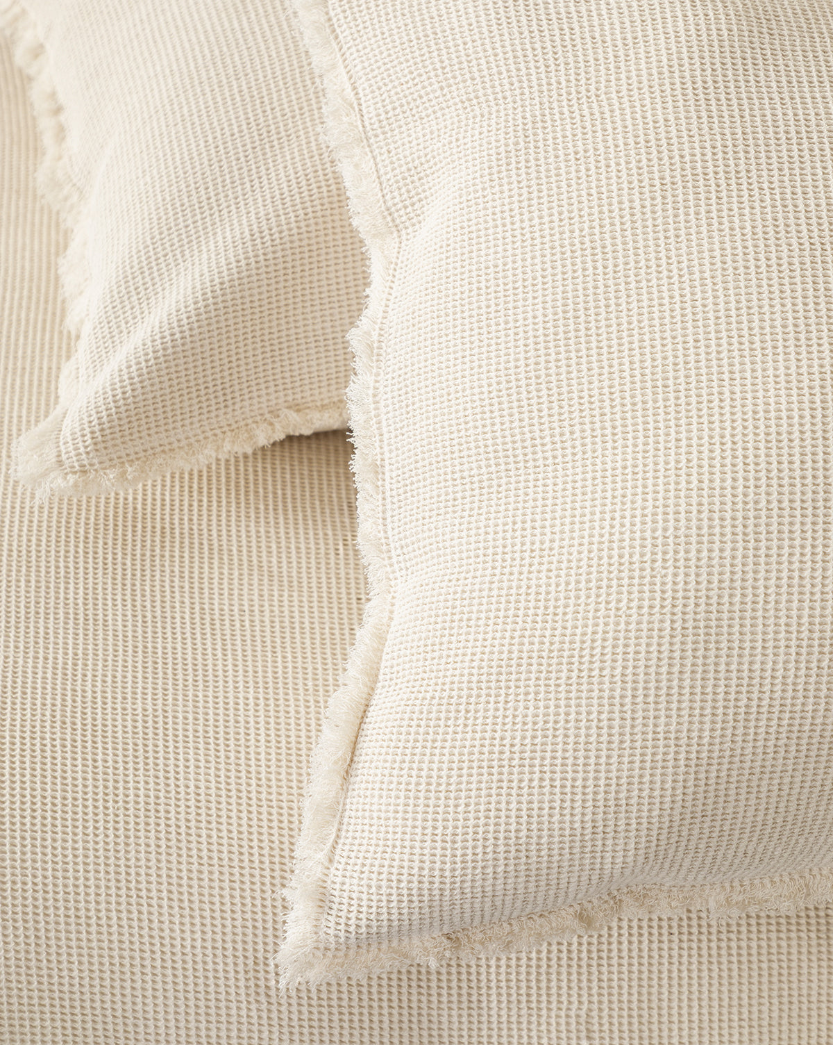 Close-up of the Helene Waffle Shams (Set of 2), featuring cream-colored cotton waffle weave fabric and fringed edges, displayed on a matching neutral surface.