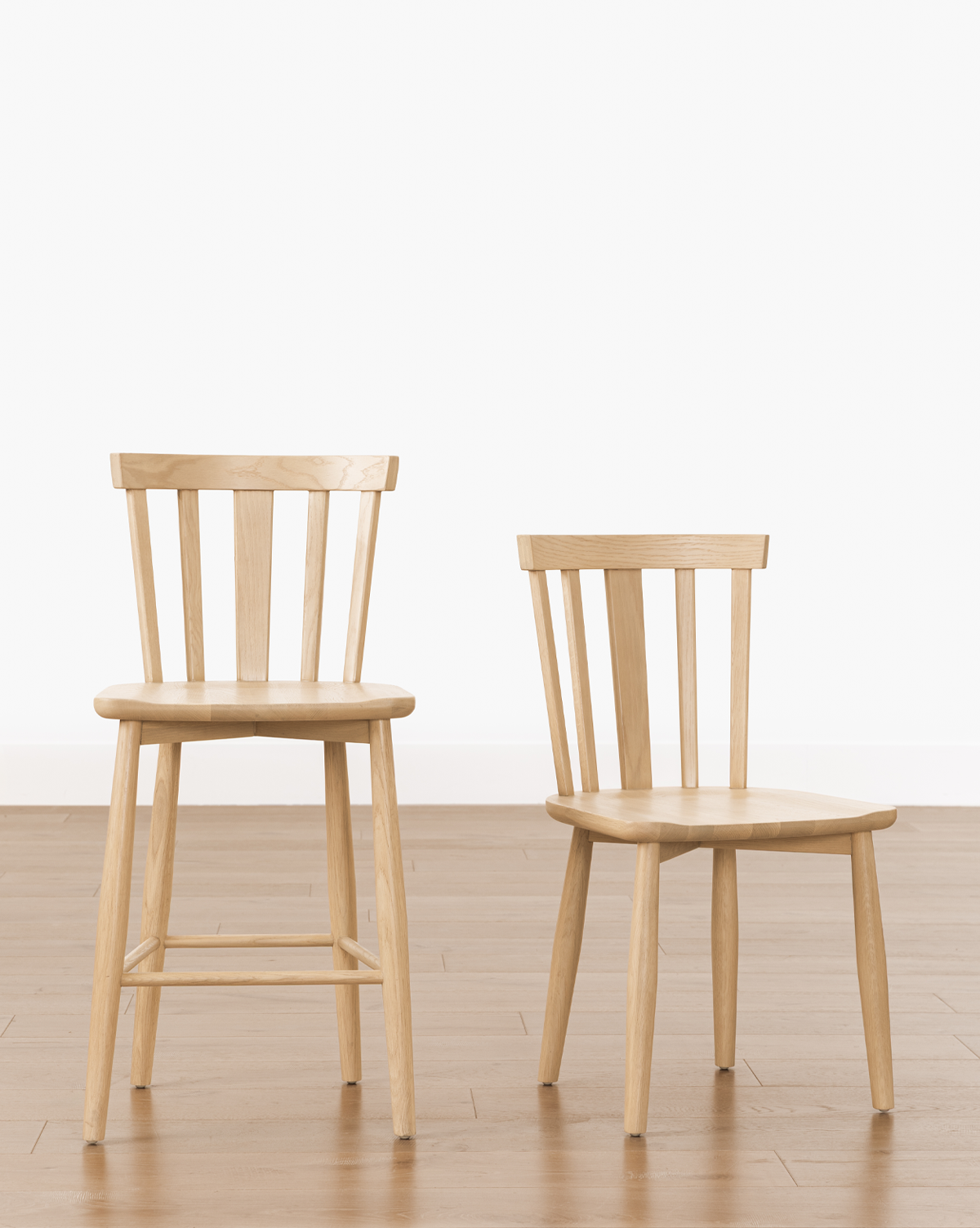 Two Hemingway Chairs—one taller with a footrest and one shorter—both with vertical slatted backs, are placed side by side on a wooden floor against a plain white background, by McGee & Co.