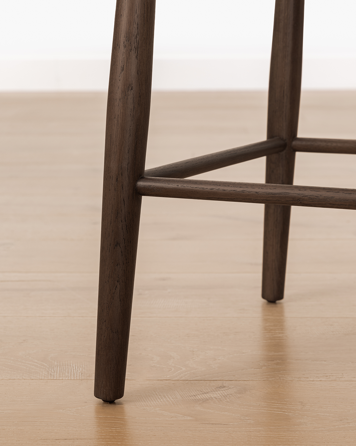Close-up of the Hemingway Chair's wooden legs and footrest on a light wood floor, highlighting its dark brown finish and sturdy, simple design—this solid wood chair brings timeless appeal to any space, by McGee & Co.