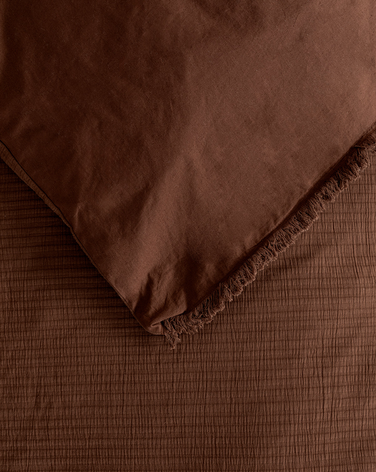 A close-up of the Hendricks Chocolate Cotton Duvet Cover with fringed edge draped over a textured brown surface in matching tones, creating a simple, cozy look.