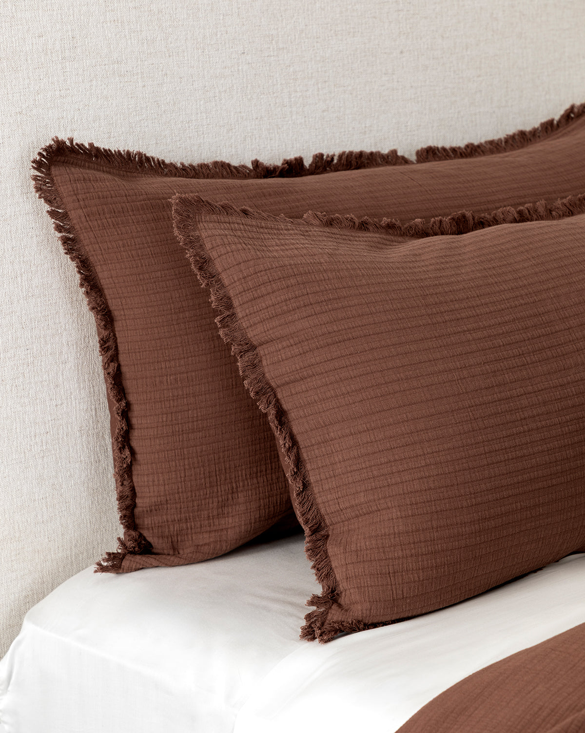 Two Hendricks Chocolate Cotton Shams (Set of 2) rest against a light fabric headboard on a white bed. A matching brown 100% cotton bedding or sheet is partially visible at the bottom.