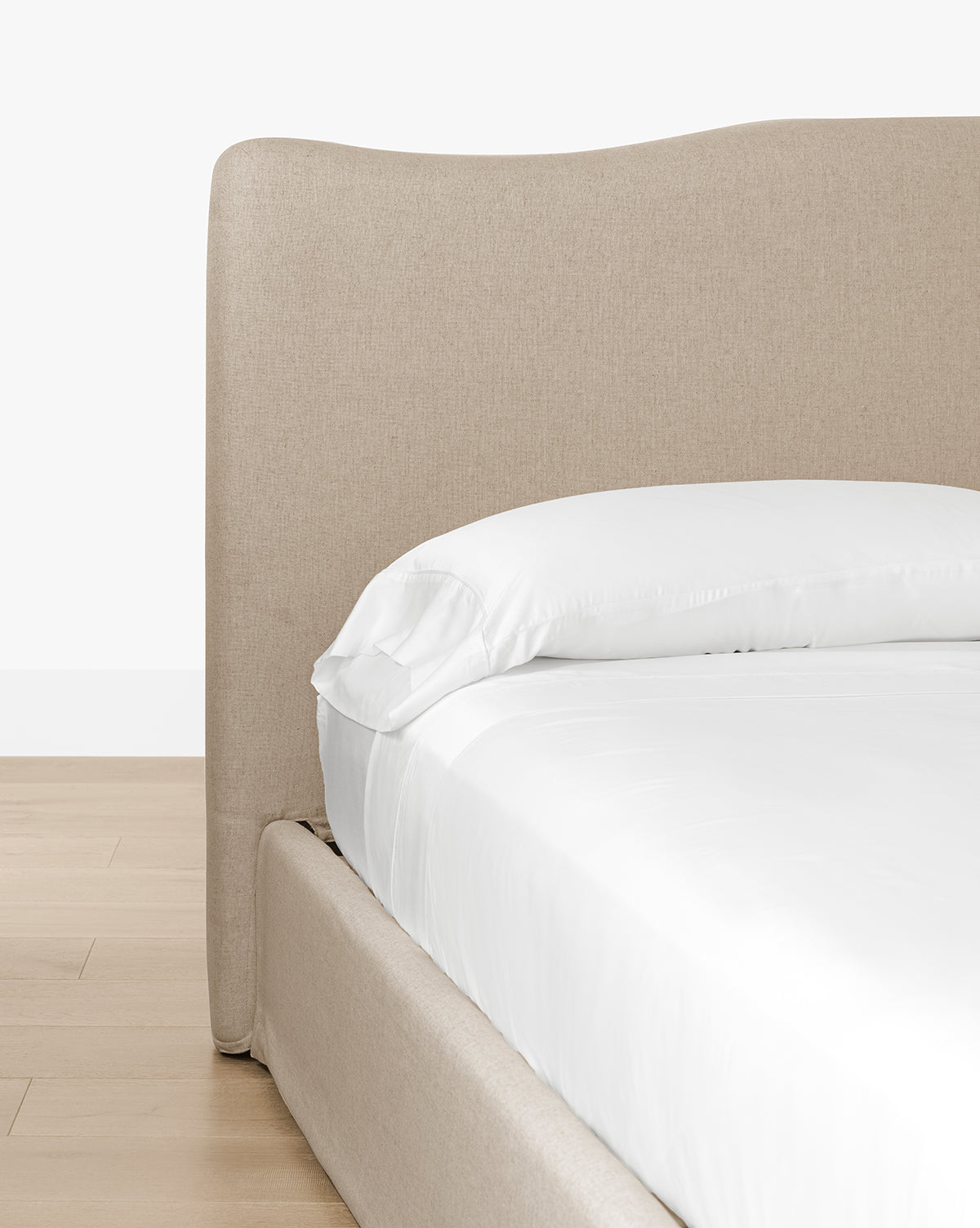 A close-up of the Henrietta Bed (Ready to Ship) features a beige upholstered frame with a tall, curved headboard, styled with white sheets and pillow on a light wood floor against a white wall for timeless bedroom appeal, by McGee & Co.