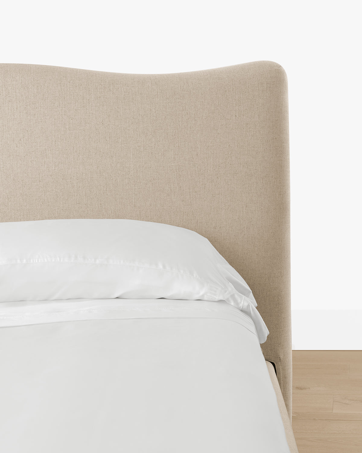 A close-up of the Henrietta Bed (Ready to Ship) featuring a light beige curved headboard, white pillow, and sheets on a light wood floor against a plain white background—perfect for modern bedroom styling, by McGee & Co.