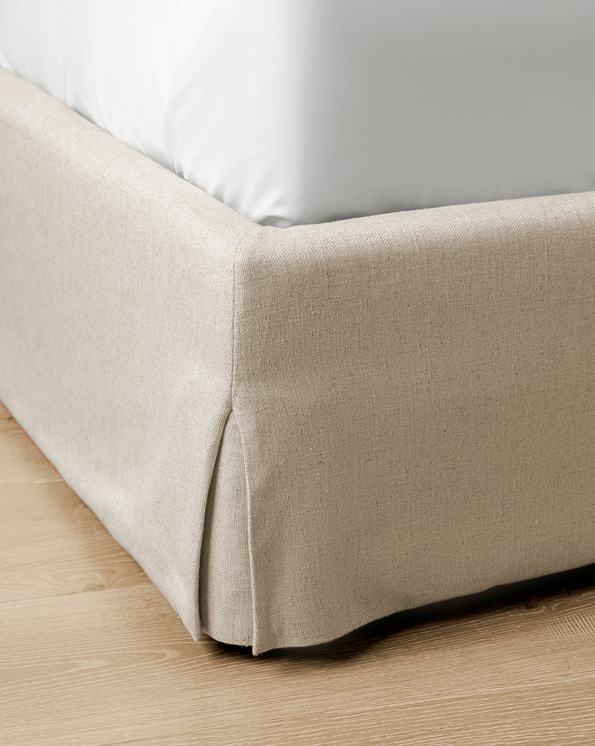 Close-up of the Henrietta Bed (Ready to Ship), showcasing its beige upholstered base with a neat corner pleat and a white mattress, set on a light wooden floor—an ideal choice for stylish bedroom updates, by McGee & Co.