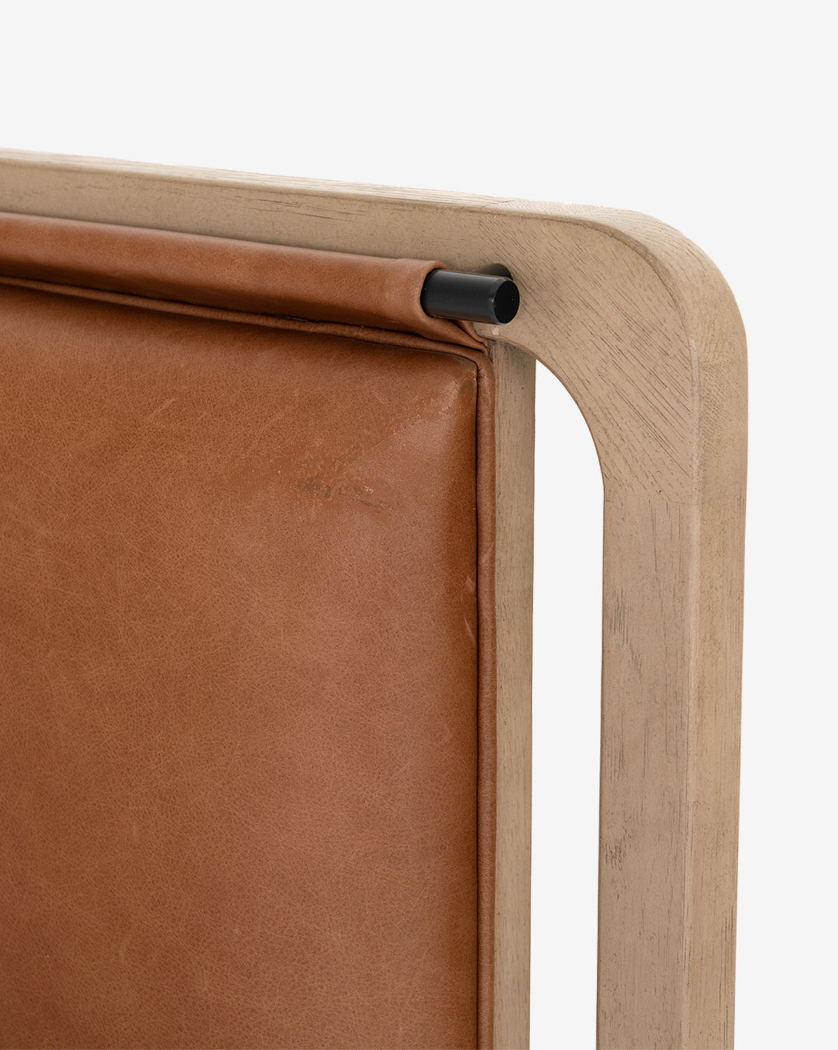 Close-up of the Hensley Bed: a wooden frame with a tan leather headboard cushion, attached by a black metal rod. The corner joint and upholstery detail highlight modern design, set against a white background.