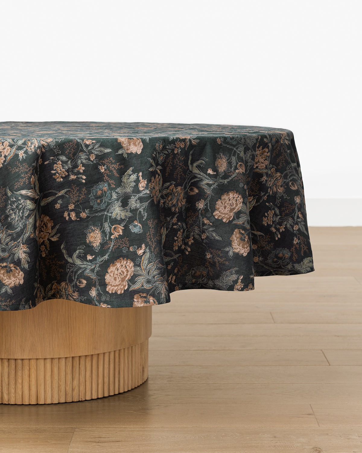 A round table with a light wood base is draped in the Hestercombe Tablecloth, showcasing vintage-inspired muted pink and green florals. The table sits on a light wooden floor against a plain white wall, by McGee & Co.
