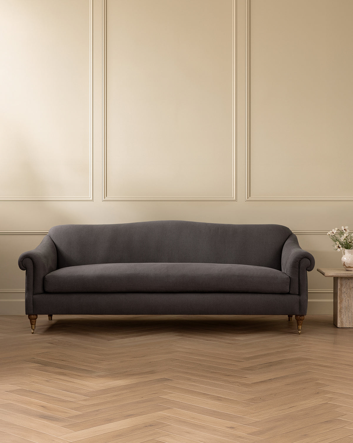 A dark gray Hildegard Sofa 90 with rolled arms sits against a beige paneled wall on a herringbone wood floor. To its right, a small stone side table holds a vase of flowers next to the handcrafted sofa, by McGee & Co.