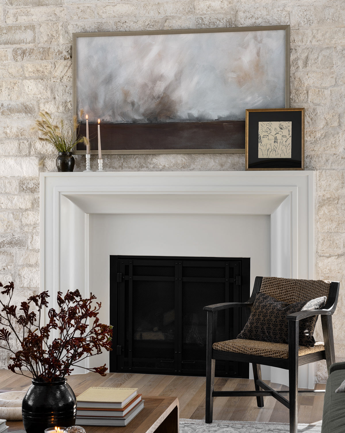 A modern living room styled with the Courtyard fireplace, abstract art and vintage illustration above, candles, a vase with grasses, a patterned black-framed chair, and a coffee table set with books and dark red foliage.