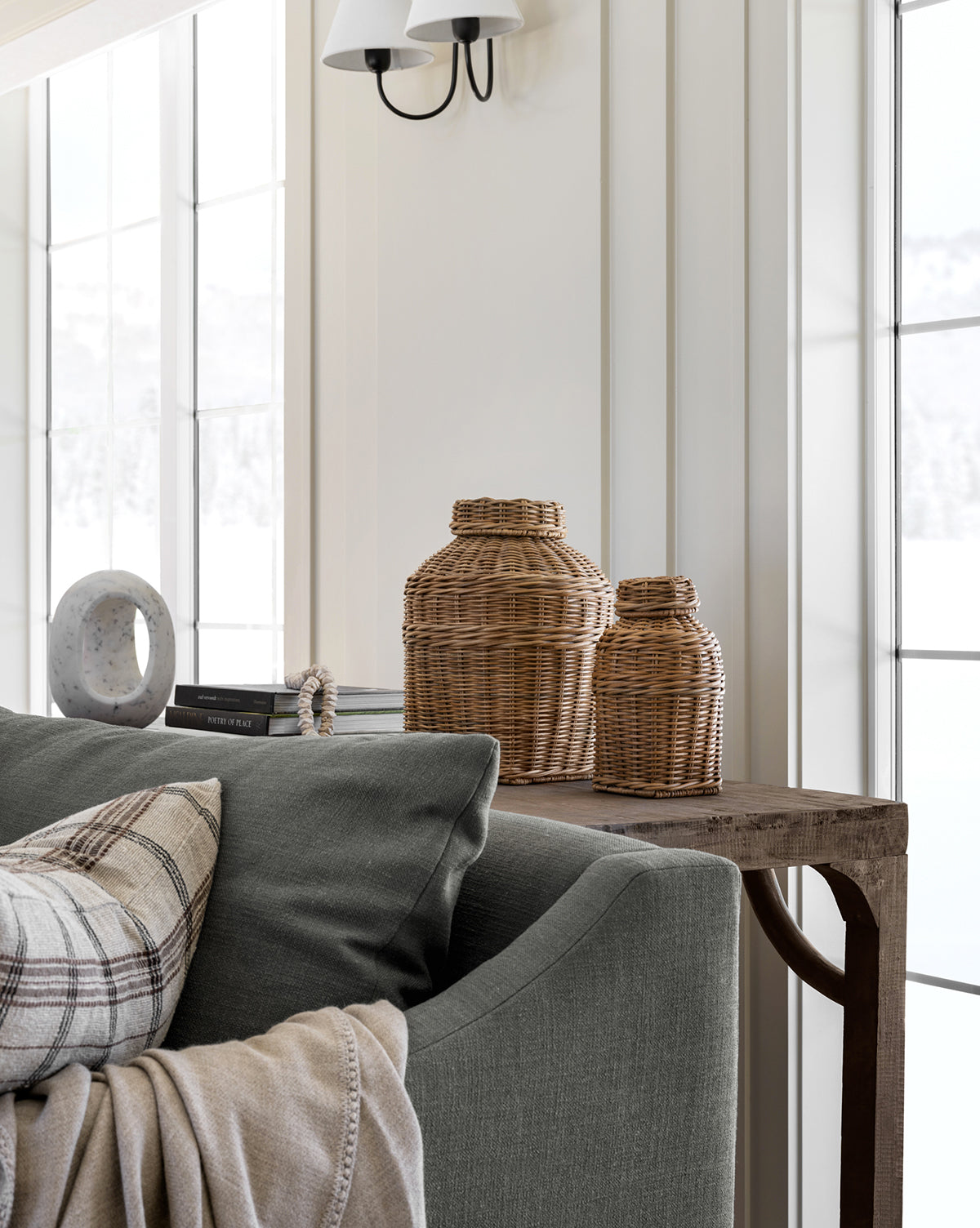 A cozy living room corner features a gray sofa, plaid pillow, beige throw, and rustic wood table topped with two Ahnamia Wicker Jars, stacked books, and a round stone decor piece by tall windows with white trim.