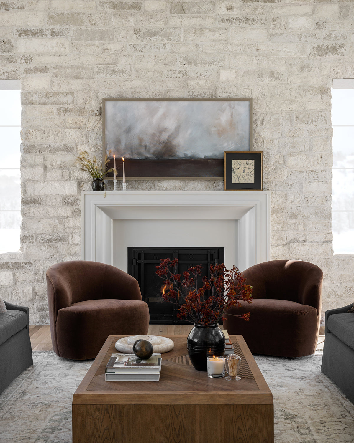 Modern living room with light stone walls, a white fireplace, two brown velvet armchairs, a wooden coffee table with décor, and Stormy Expanse artwork displayed in a frame above the mantel alongside candles.