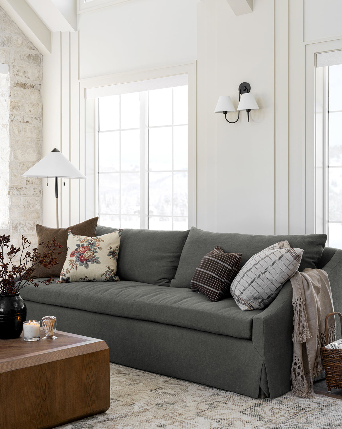 A cozy living room featuring the Community Everleigh Slipcover Sofa 96.5 in dark gray, styled with patterned throw pillows, a wooden coffee table with candles, large sunlit windows, and neutral walls and flooring, by McGee & Co.