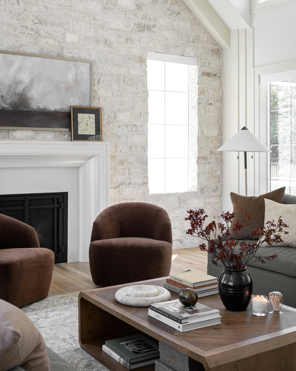 A modern living room features a stone accent wall, brown armchairs, and a wooden coffee table displaying books, a vase of red foliage, and the Jackson Offset Sphere. A white fireplace with artwork shines as natural light streams in.