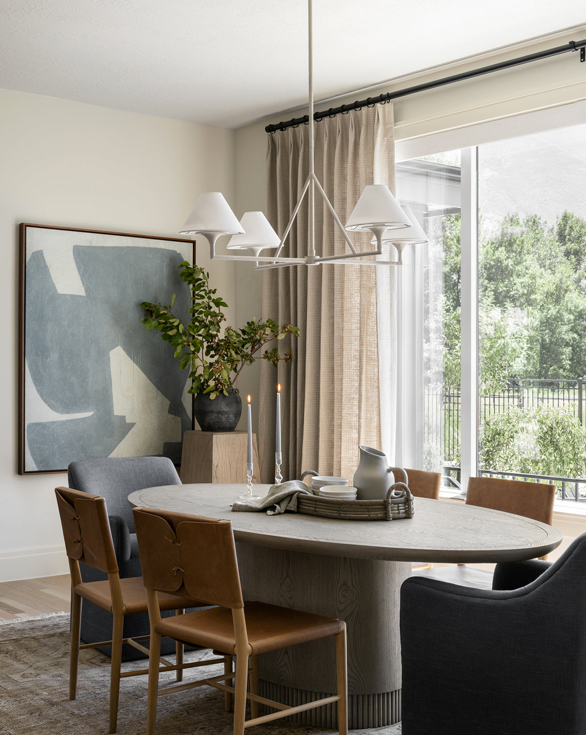 A modern dining room features a round wooden table, Danson Dining Chairs with leather seats, abstract wall art, candles, a vase of branches, decorative books, and large windows with beige curtains overlooking greenery by McGee & Co.