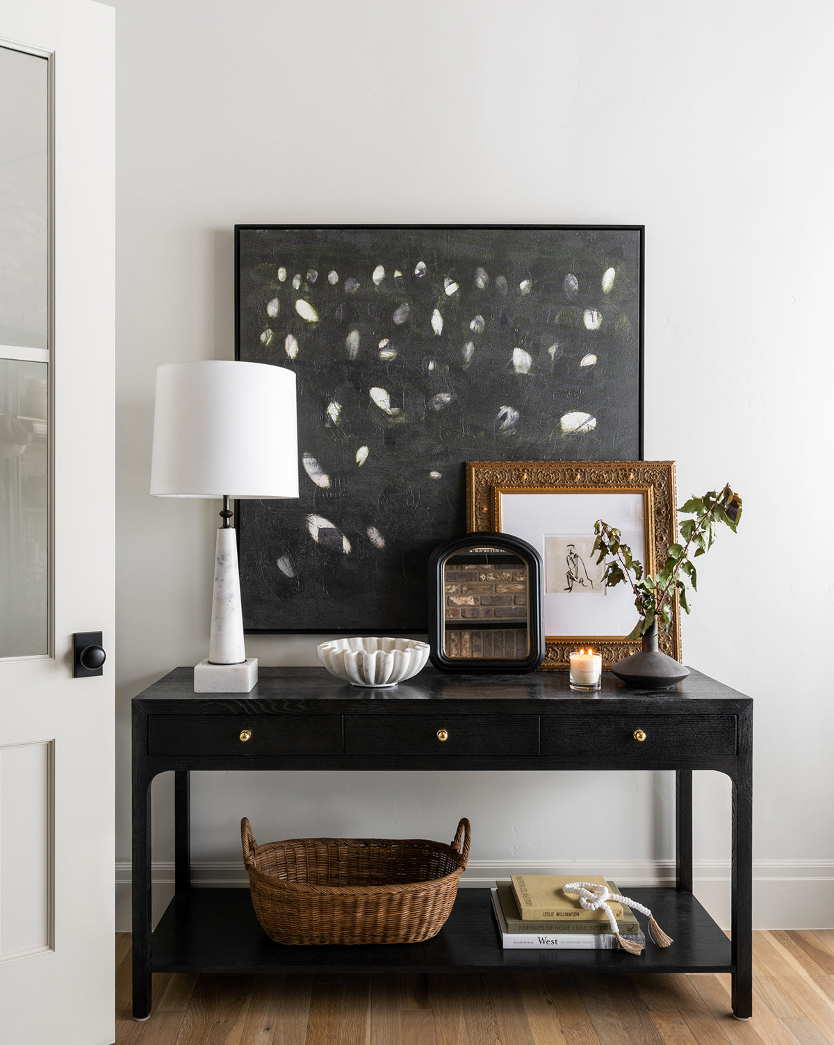 A black console table against a white wall displays a marble lamp, decorative bowl, framed art, candle, leafy vase, and small cabinet. A wicker basket sits below. Above hangs Verde Oculus by Jamie Beckwith in a black maple floater frame.