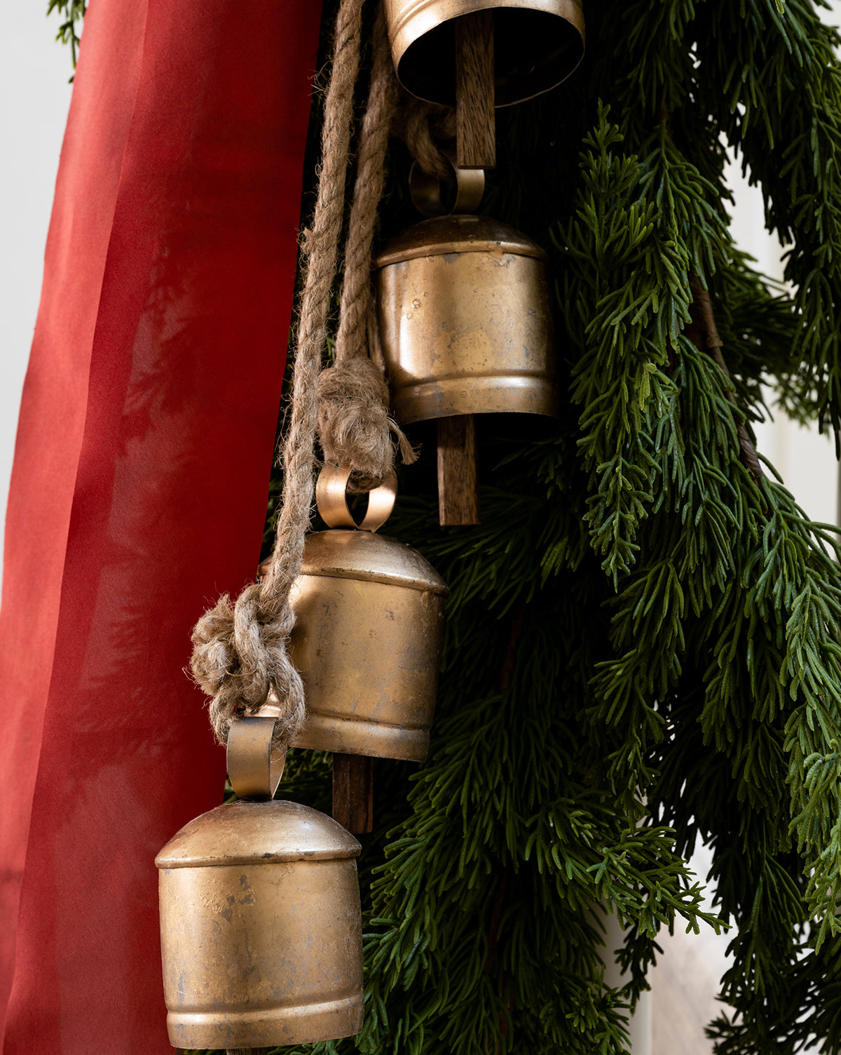 Close-up of three Hanging Iron Bells with a gold finish, suspended by thick rope beside green fir branches and a red ribbon, adding festive charm and rustic holiday décor, by McGee & Co.