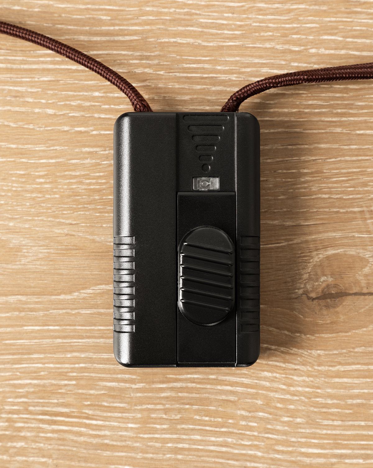 The Holloway Floor Lamp’s black inline switch, featuring a ridged sliding button and attached to a brown fabric-covered cord, sits on a light wood surface, by McGee & Co.