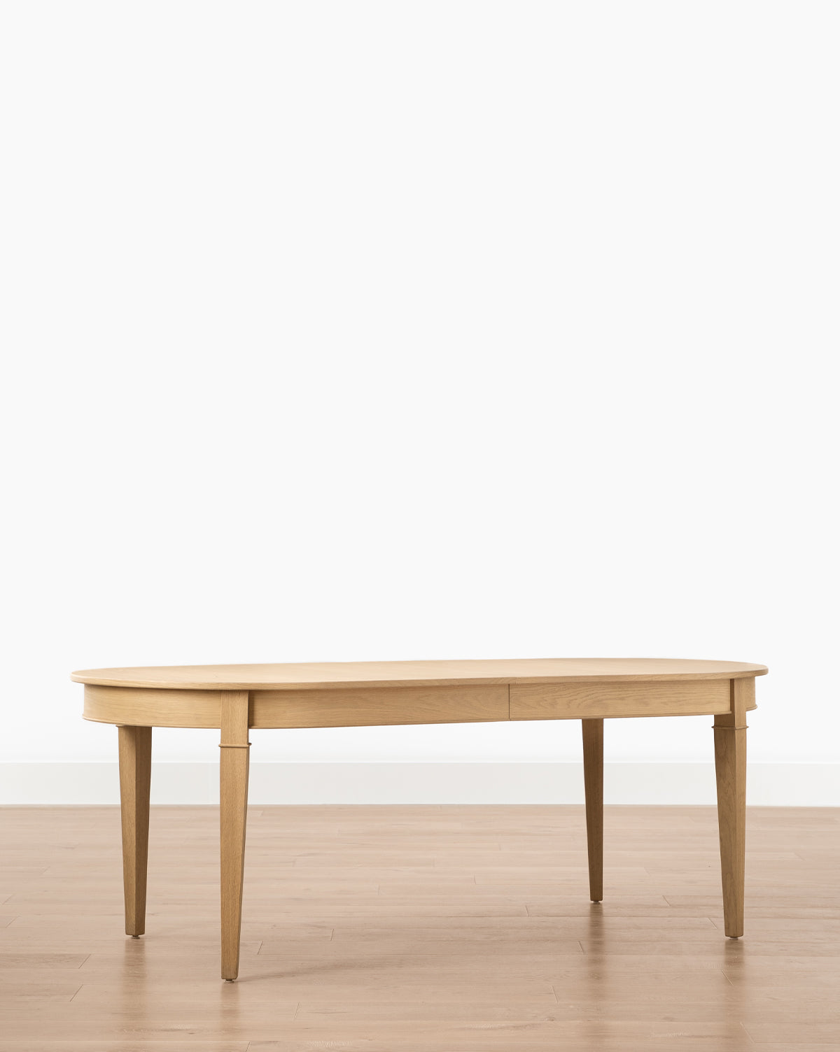 The Hughes Oak Extension Dining Table, featuring an oval shape and four tapered legs, is displayed on a light-colored wooden floor against a plain white background by McGee & Co.