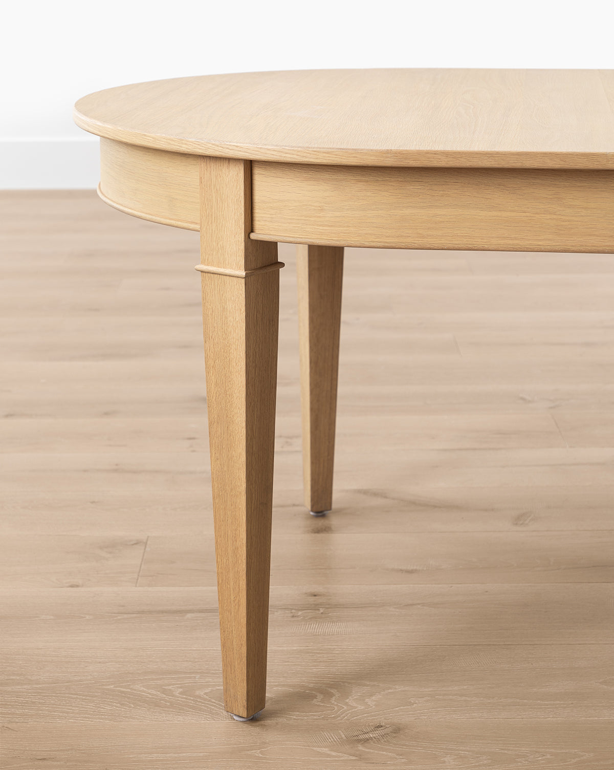 Close-up of the Hughes Oak Extension Dining Table, an oval table with a self-storing butterfly leaf and tapered legs, set on a matching light wood floor against a white wall, by McGee & Co.