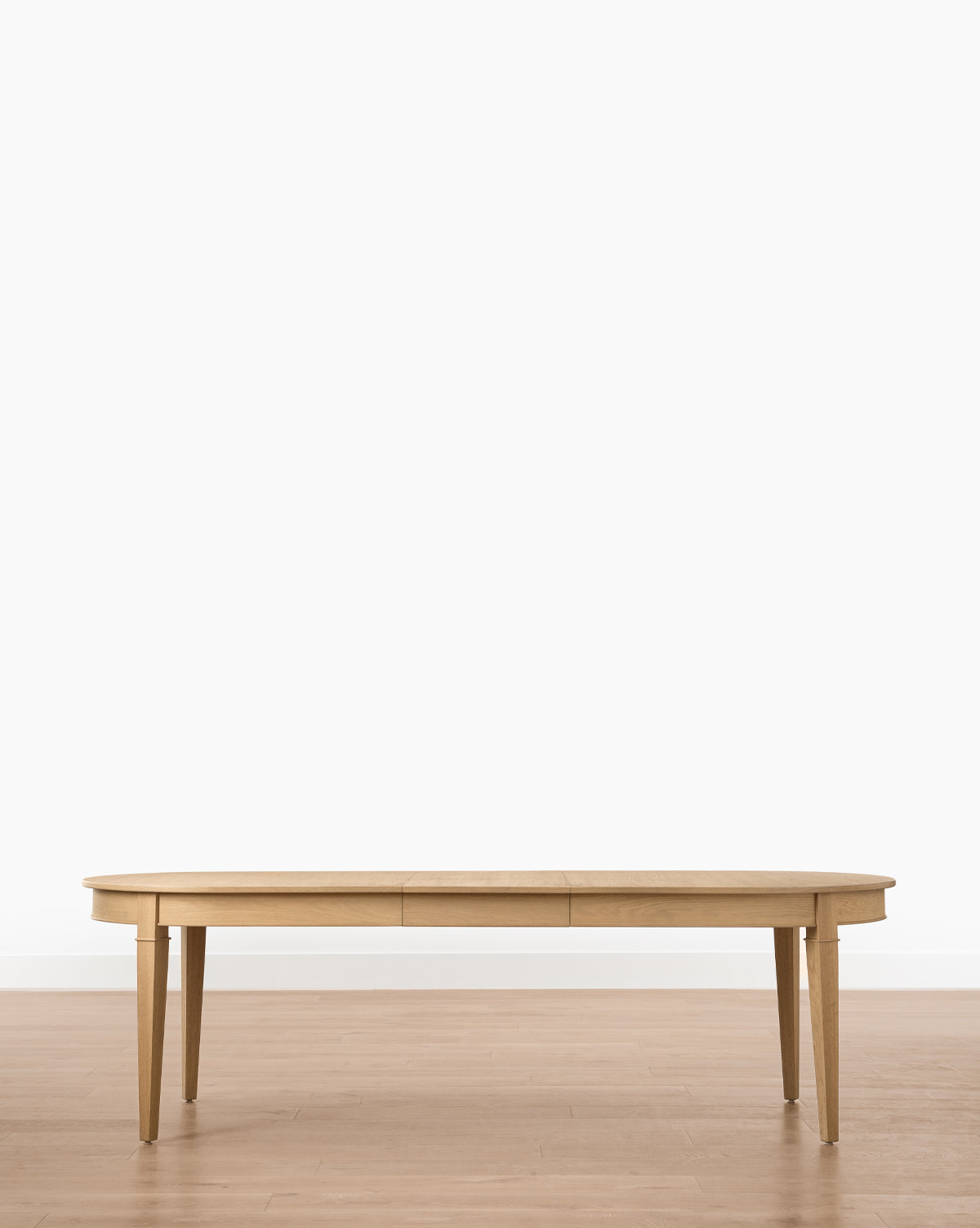 The Hughes Oak Extension Dining Table, featuring tapered legs and a self-storing butterfly leaf, stands on a light wood floor against a plain white wall for versatile dining, by McGee & Co.