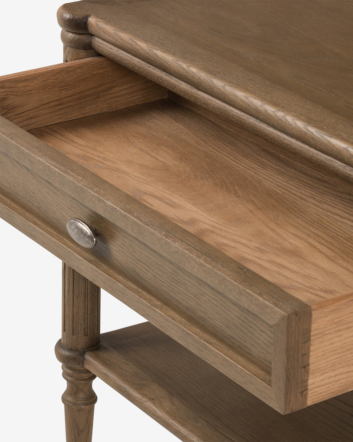 A close-up of the Humphrey Nightstand in oak shows its open drawer with a round metal knob and elegant turned legs. The natural wood finish adds charm, making this nightstand a timeless addition to any traditional bedroom décor, by McGee & Co.