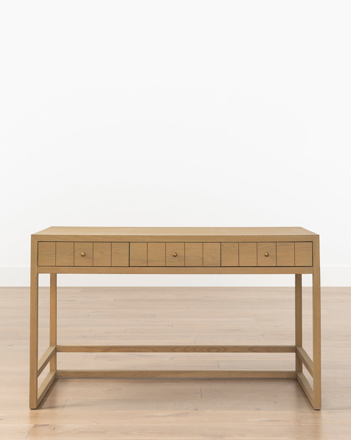 The Huxton Desk features a light finish, rectangular shape, and clean lines, with four small drawers with brass pulls. Its minimalist design pairs nicely with a matching wooden floor and plain white wall, by McGee & Co.
