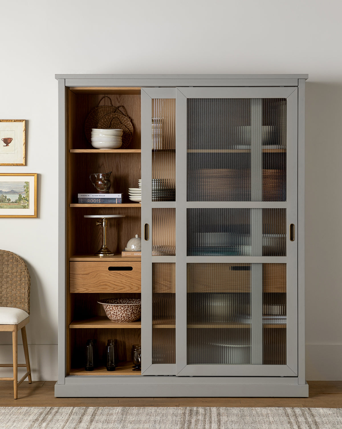 The Phillips Cabinet, featuring reeded glass doors, antique brass handles, and open shelves for dishes, bowls, books, and decor, stands against a white wall beside a woven chair by McGee & Co.