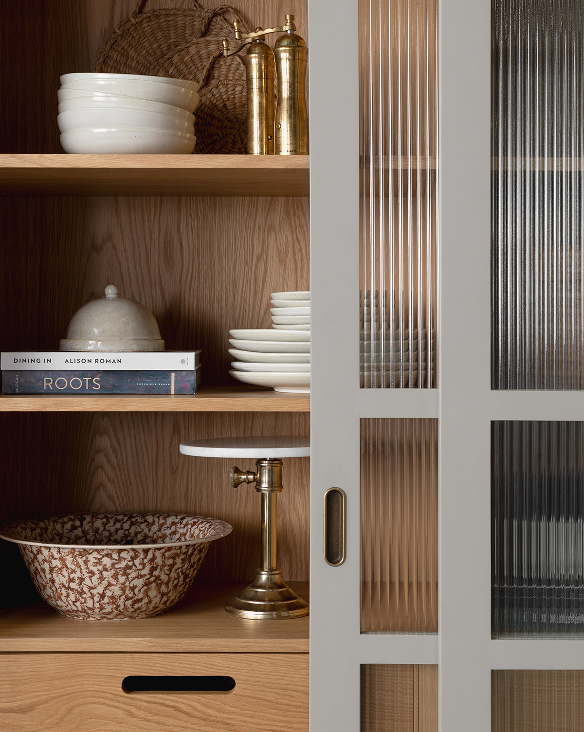 A wooden kitchen shelf, partly hidden by vertically-lined glass cabinet doors, holds stacked bowls and plates, books, a gold pepper mill and salt shaker, a lidded dish, and the Spongeware Bowl by McGee & Co.