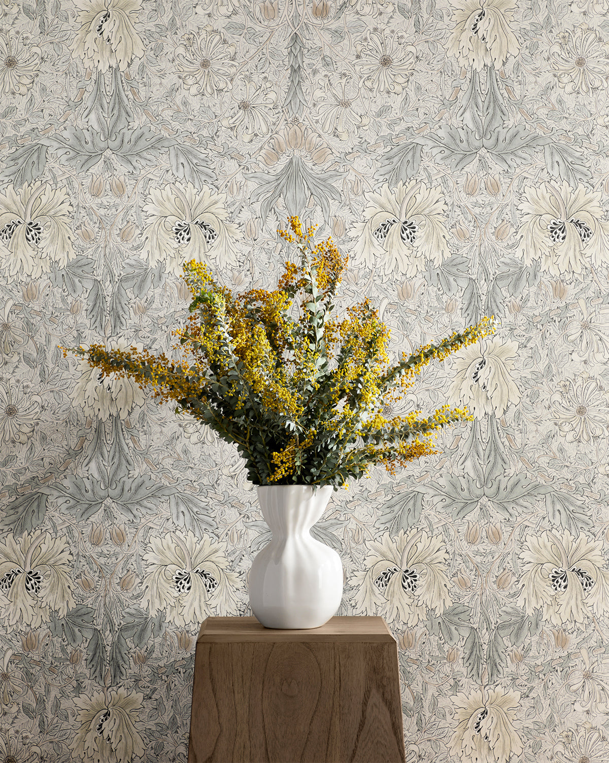 The Thessaly Vase, filled with yellow and green flowers, sits on a wooden pedestal against patterned floral wallpaper in light gray and beige tones.