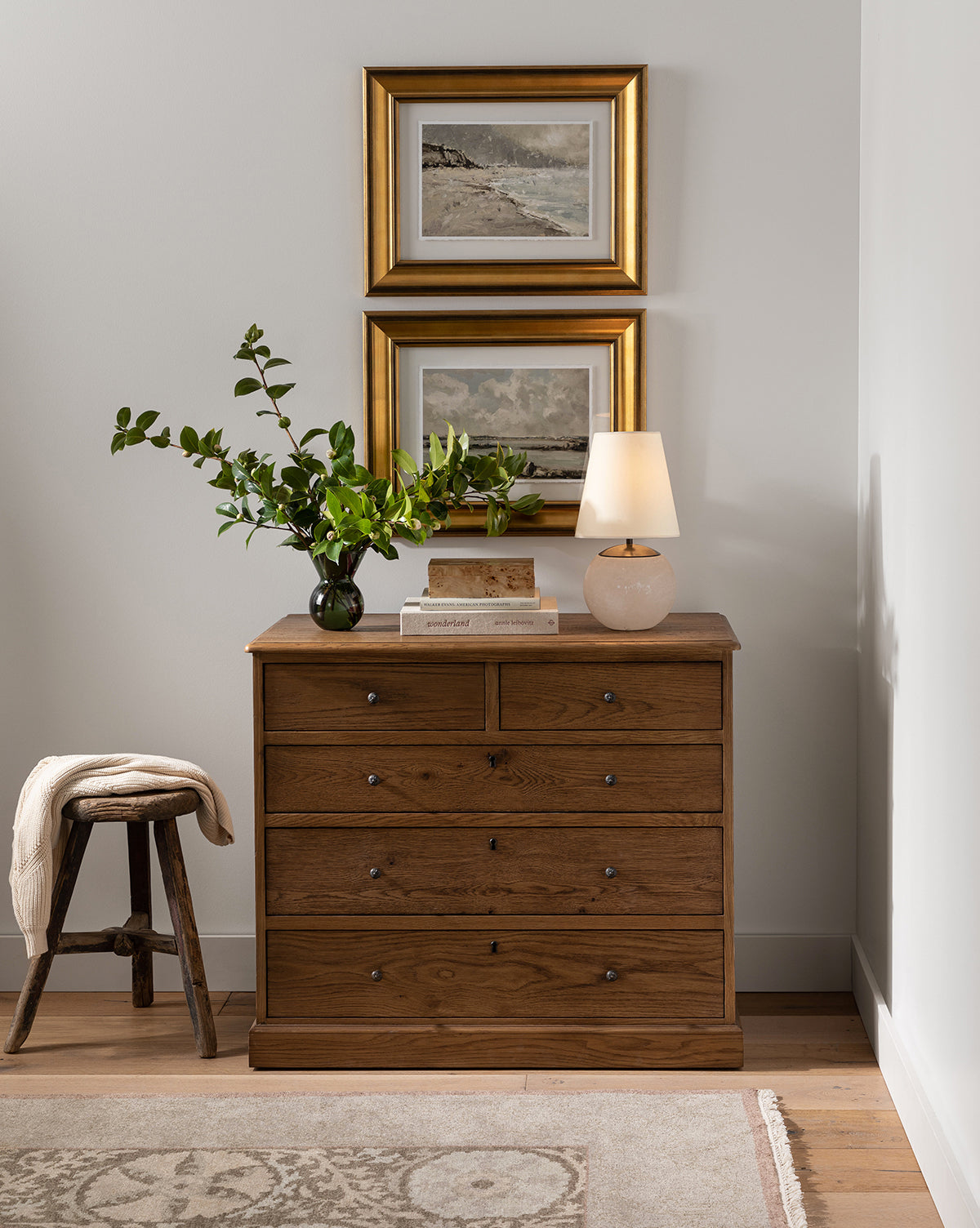 A wooden dresser with six drawers sits against a white wall, topped with a green leafy vase, stacked books, and the Terri Round Table Lamp with its alabaster stone base. Two framed landscape paintings hang above. A small wooden stool is on the left, by McGee & Co.