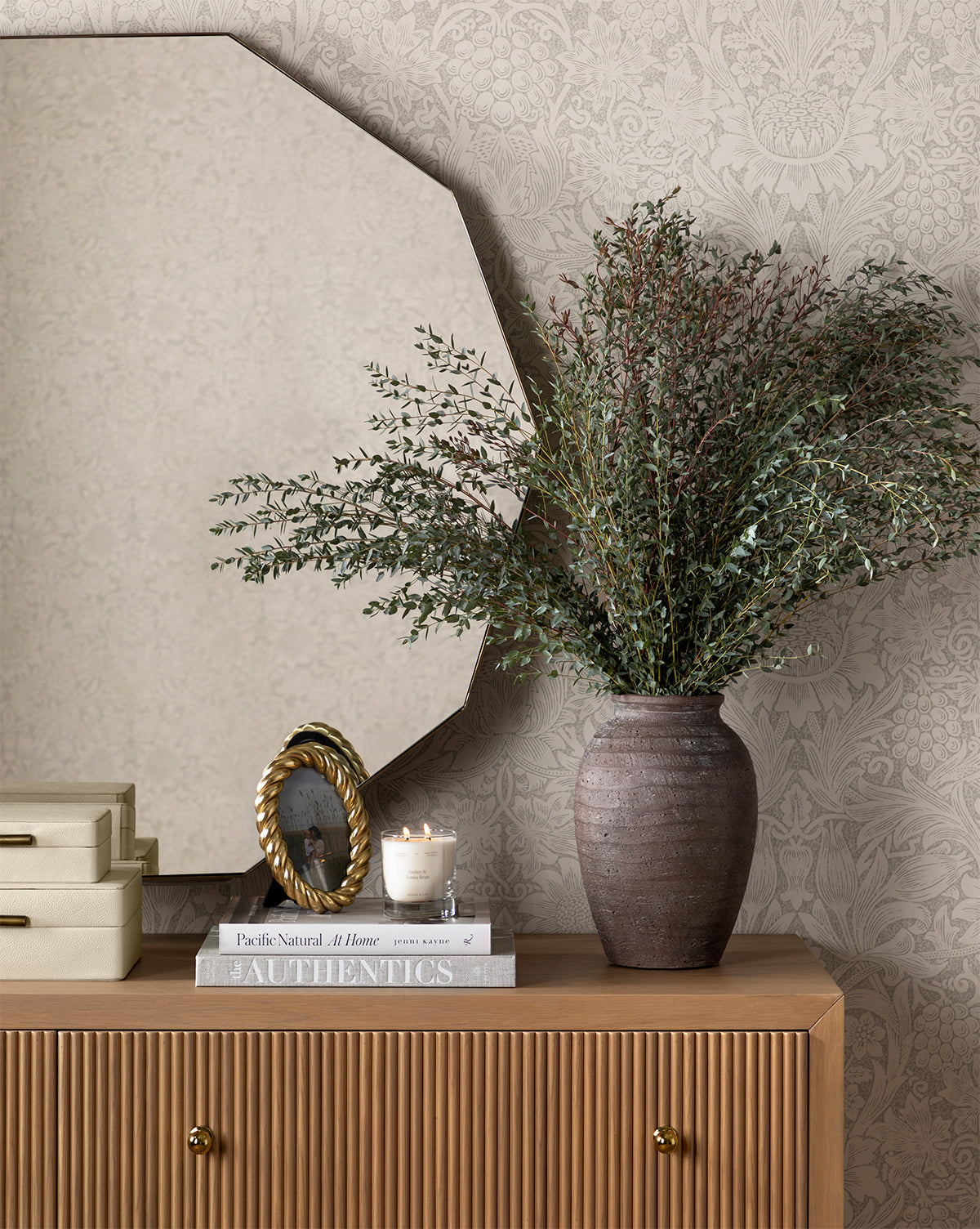 A decorative console table holds a brown vase with green foliage, stacked books, a small candle, a gold-framed photo, and two cream boxes. Above it hangs the Hawley Decagon Mirror against patterned wallpaper, by McGee & Co.