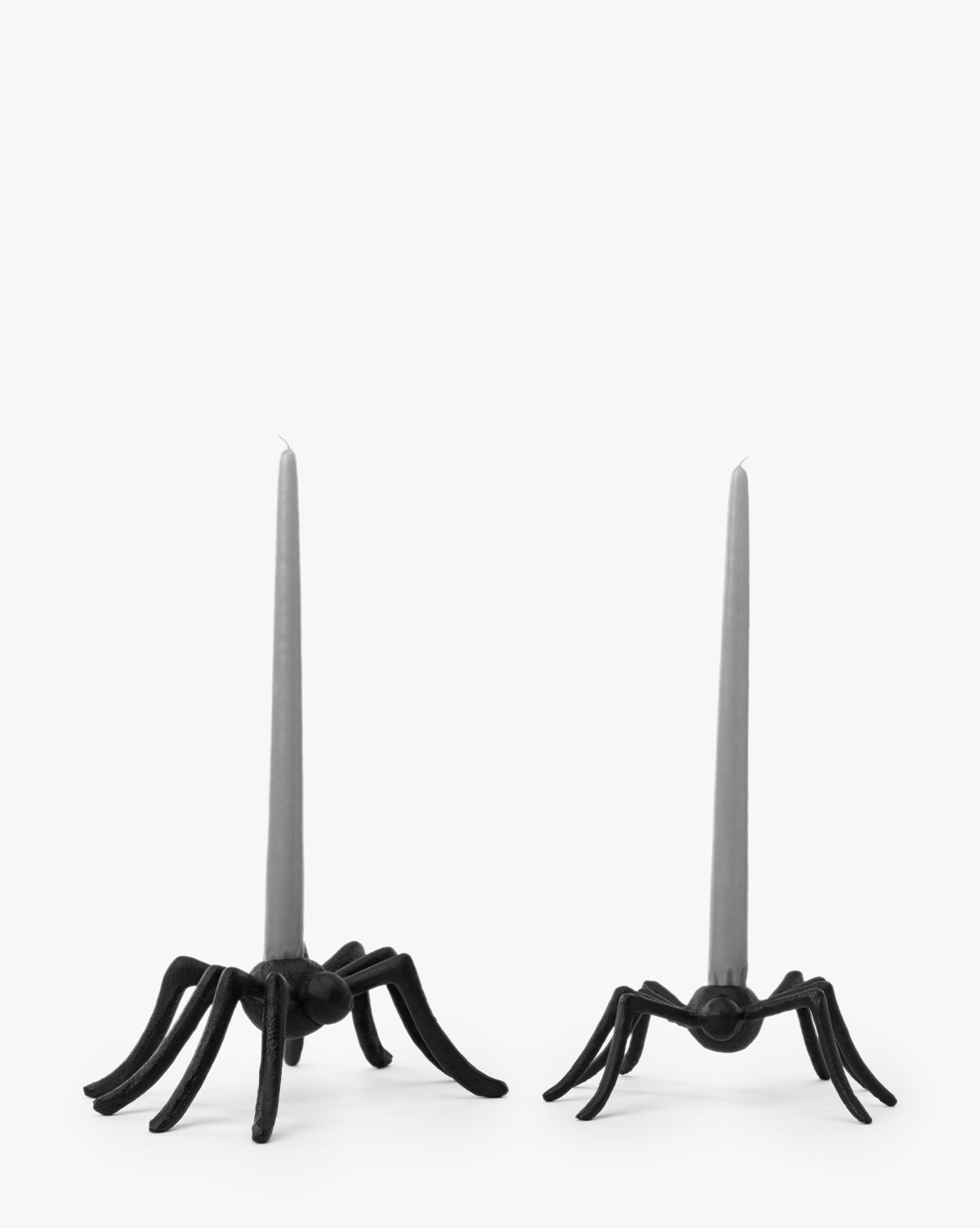 Two Iron Arachne Taper Holders, shaped like lifelike black spiders with spread legs, each hold a tall gray taper candle. These unique holders make striking Halloween decor against a plain white background, by McGee & Co.