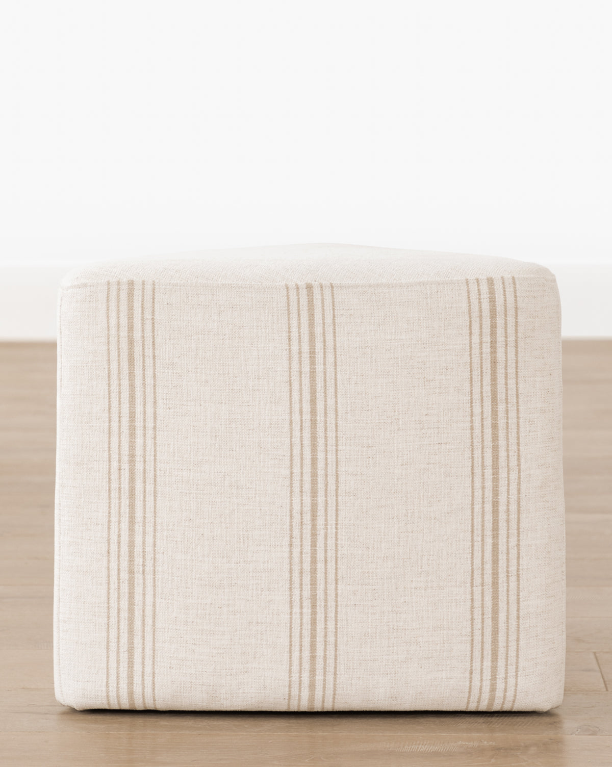 The Jacques Ottoman by Dongguan City Baby Furniture Co., Ltd features beige Crypton striped fabric in a cube shape with tan stripes—ideal for stylish, functional kids’ rooms. Shown on a wooden floor against a white wall. McGee & Co.