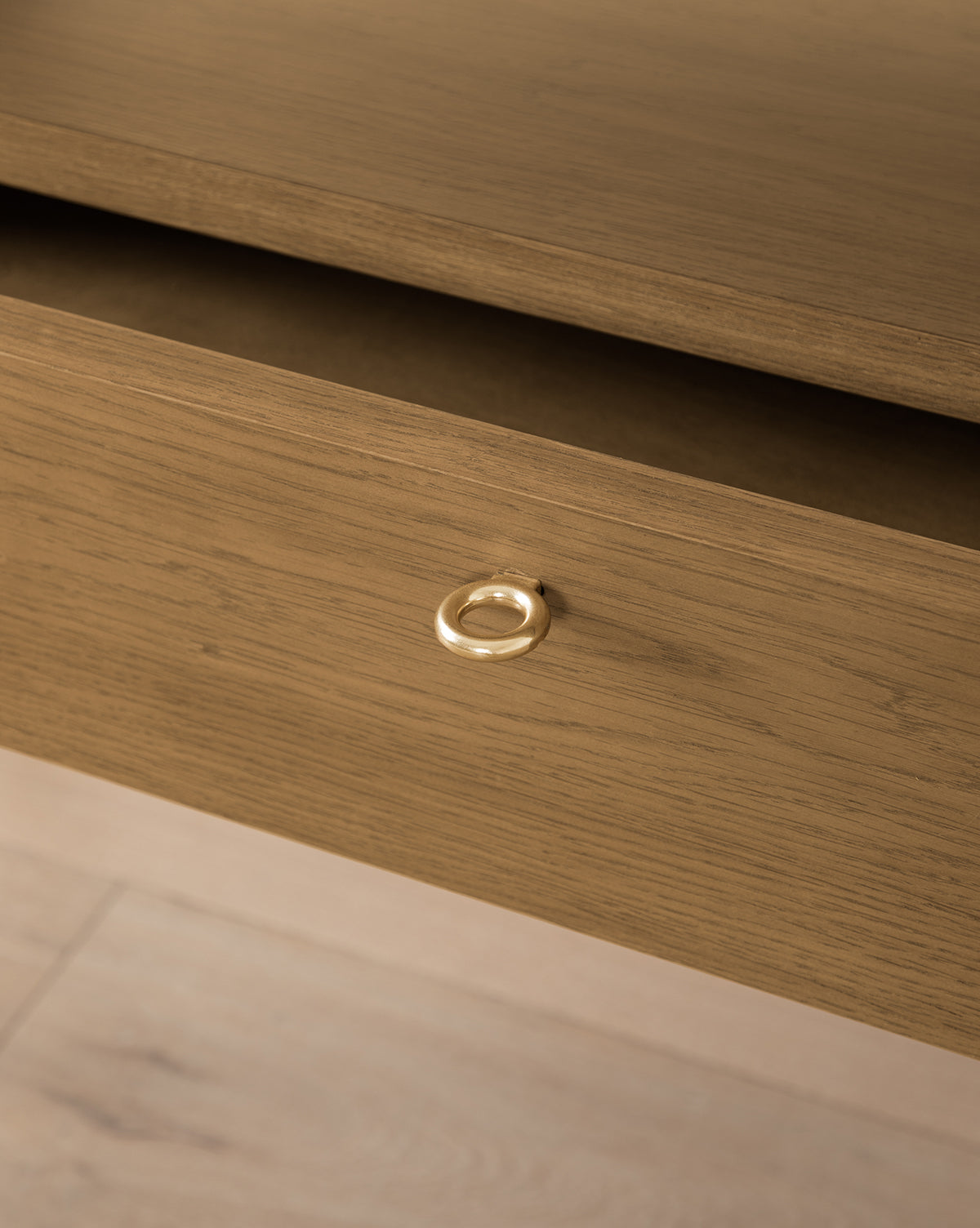 A close-up of the Jahnsen Cabinets partially open drawer with a brass ring pull highlights the honey oak finish, wood grain texture, and shows part of the light floor beneath, by McGee & Co.