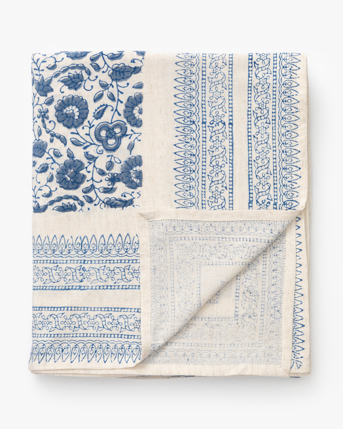 The Jennie Tablecloth is a folded cream-colored block printed tablecloth with a patchwork of intricate blue floral and geometric patterns, one corner neatly turned up to reveal the reverse side, by McGee & Co.