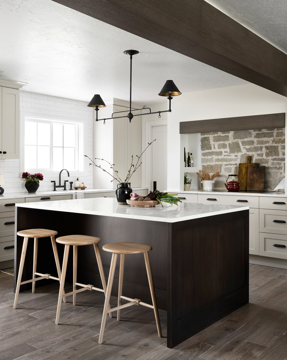 Modern kitchen featuring three Fielder Counter Stools in light oak at a large dark wood island with white countertops, neutral cabinets, a farmhouse sink, exposed brick backsplash, and minimalist pendant lights above by McGee & Co.