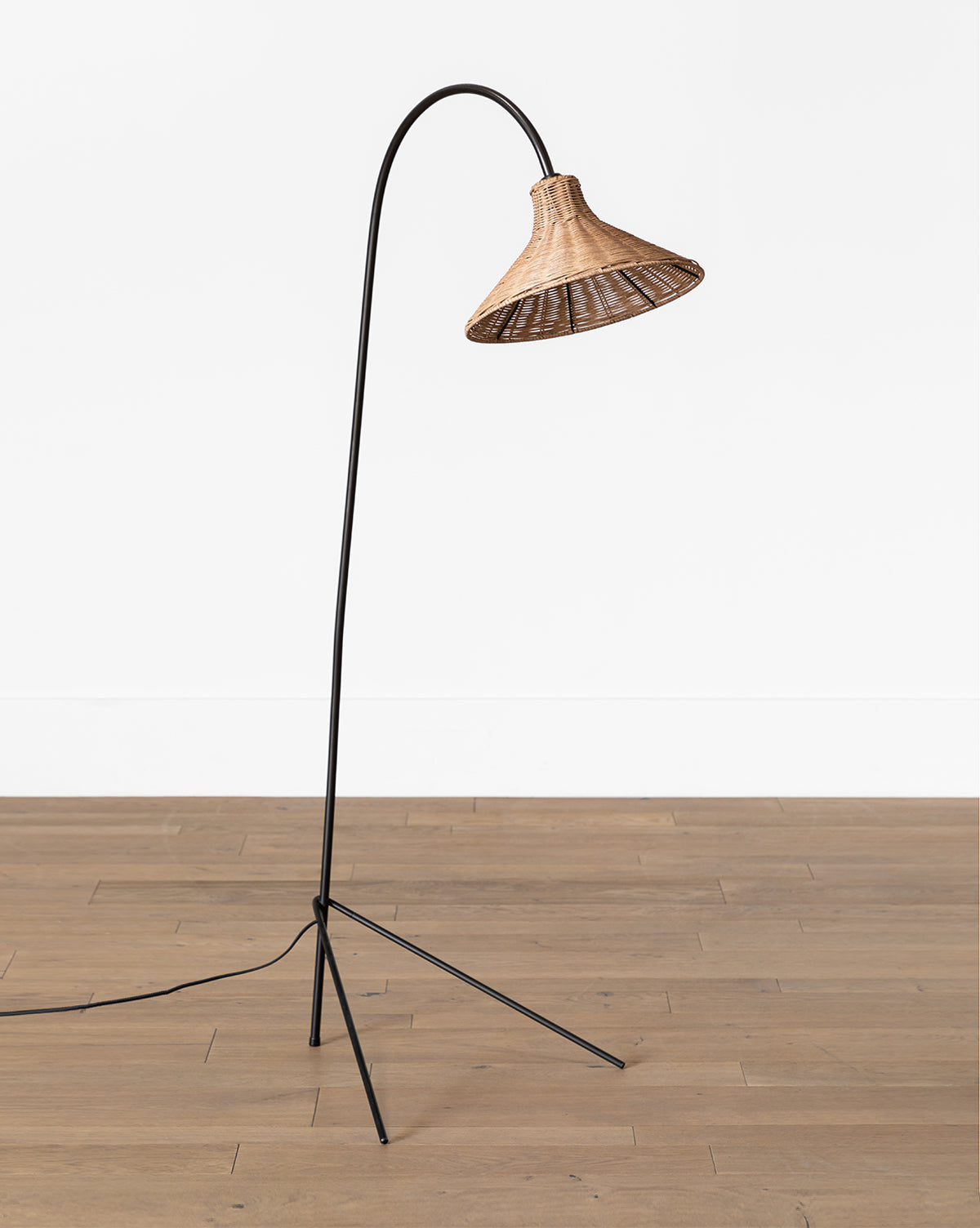 The Johanson Woven Floor Lamp by Makerspalm features a black, slender, curved metal stand and a natural rattan shade. It stands on a wooden floor against a white wall, with its cord visible—McGee & Co.