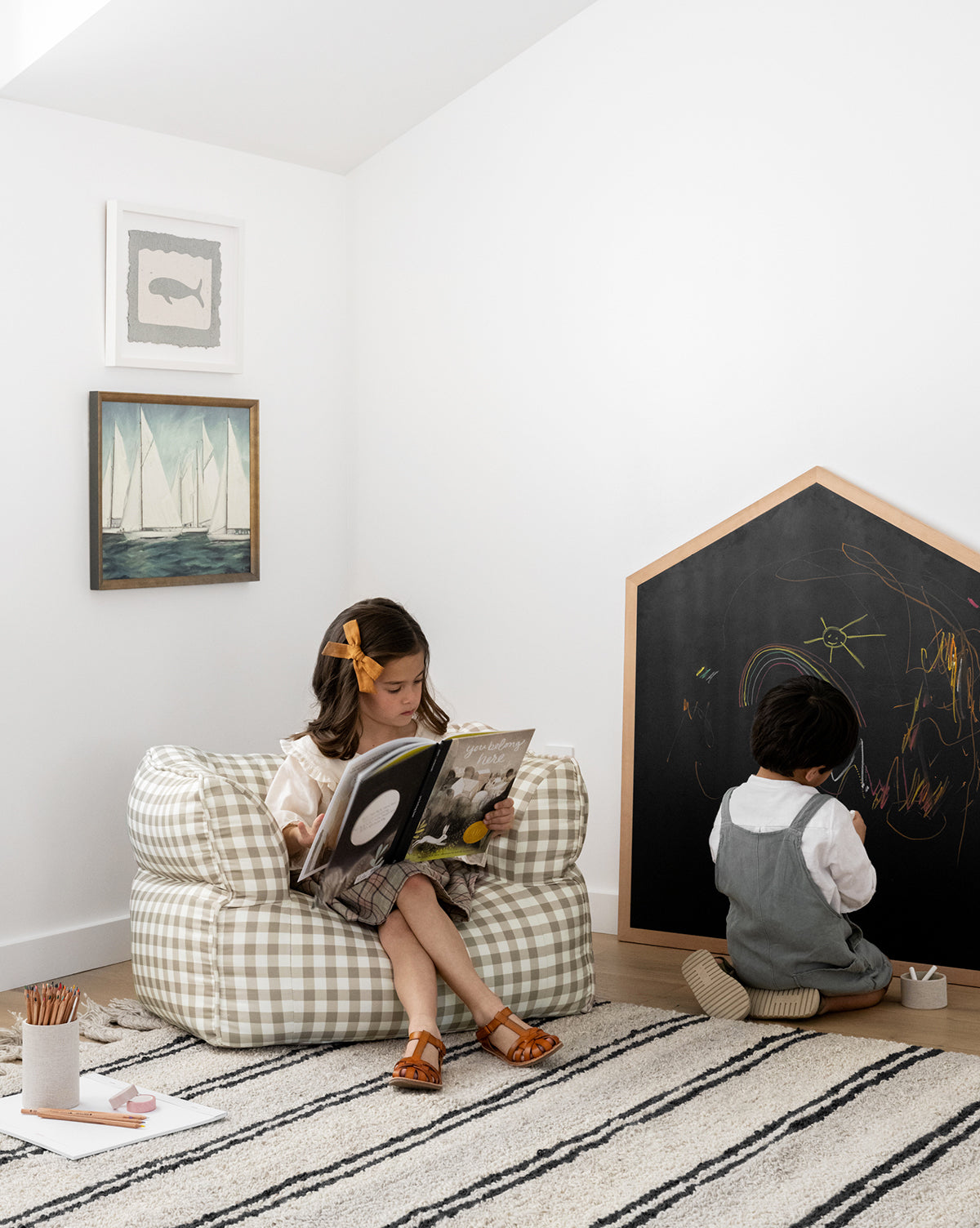 A young girl sits on a checkered bean bag reading You Belong Here, while a boy in overalls draws on a house-shaped chalkboard. Framed art and a striped rug complete the cozy, welcoming space.
