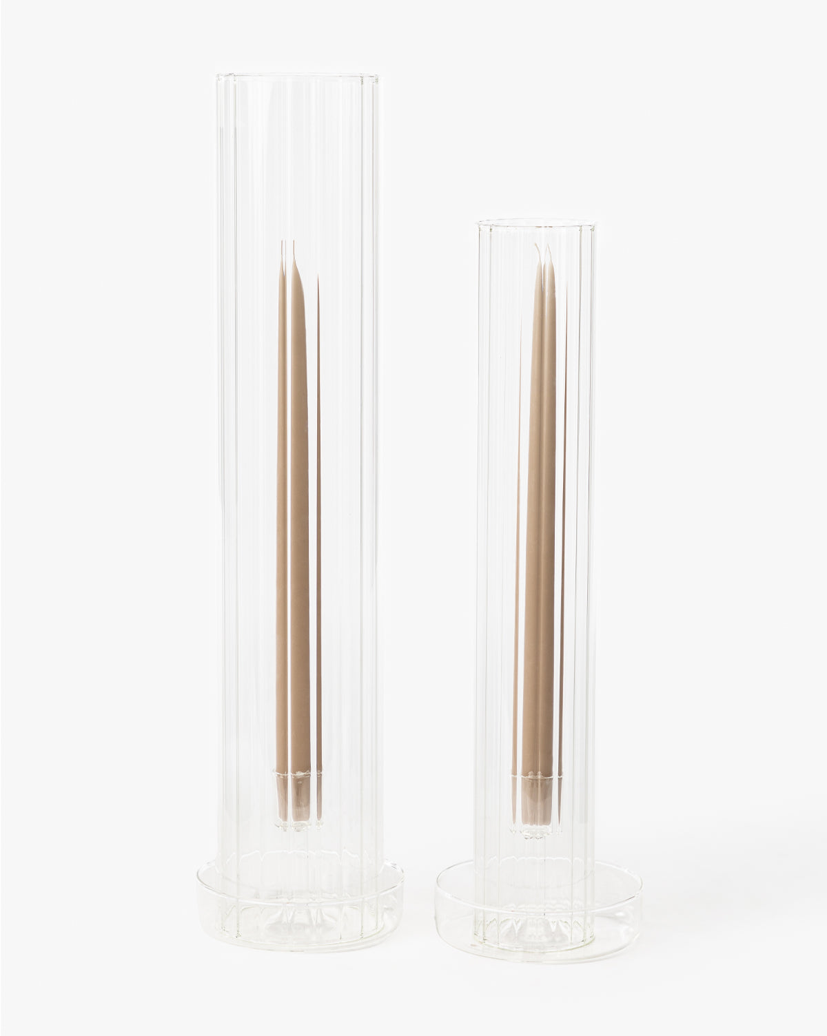 Two Kalina Ribbed Candle Sleeve holders, each tall and glass with vertical ribbing, showcase single brown taper candles. The elegant holders stand on circular bases against a plain white background.