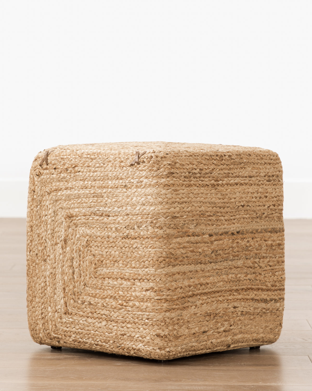 The Kelvin Woven Ottoman, hand-woven from jute, rests on a wooden floor against a plain white wall, adding versatile style to any space.